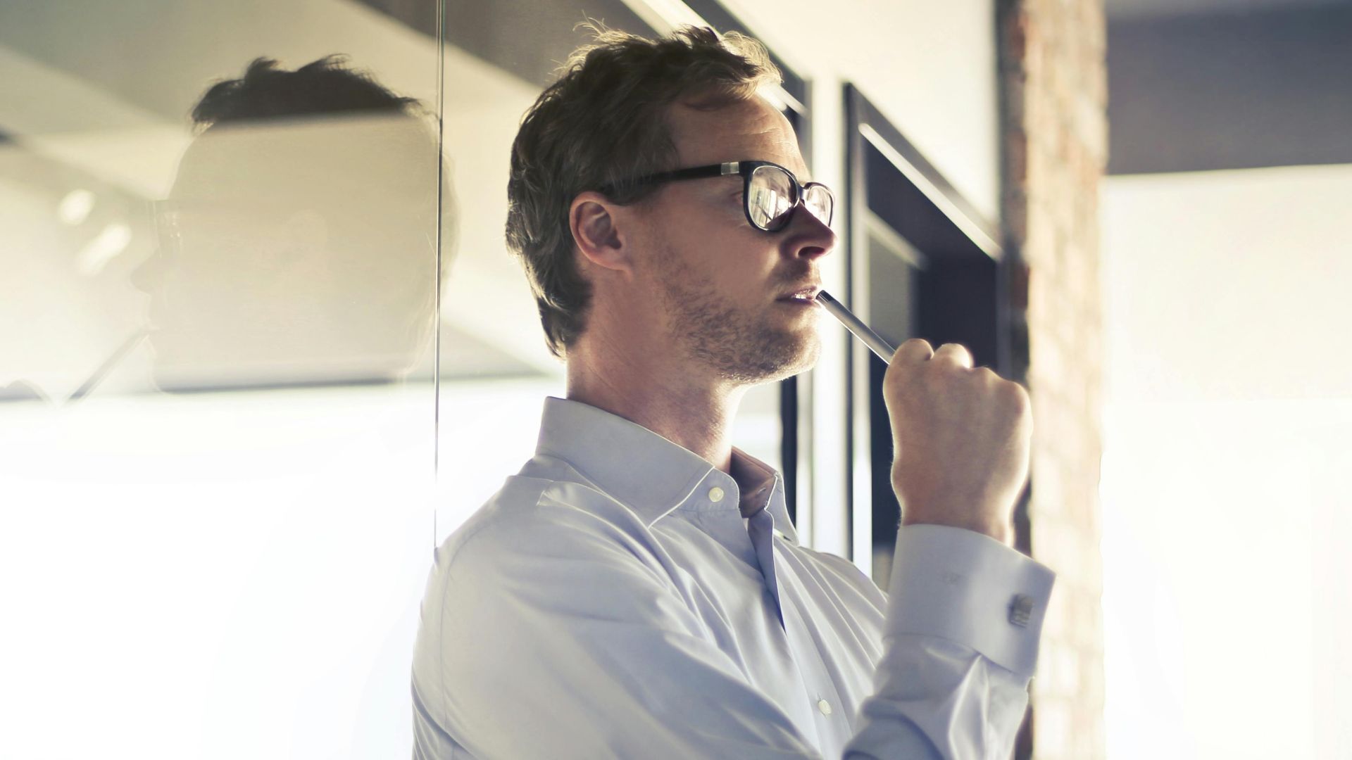 Thoughtful businessman in glasses pondering by office window, holding a folder.