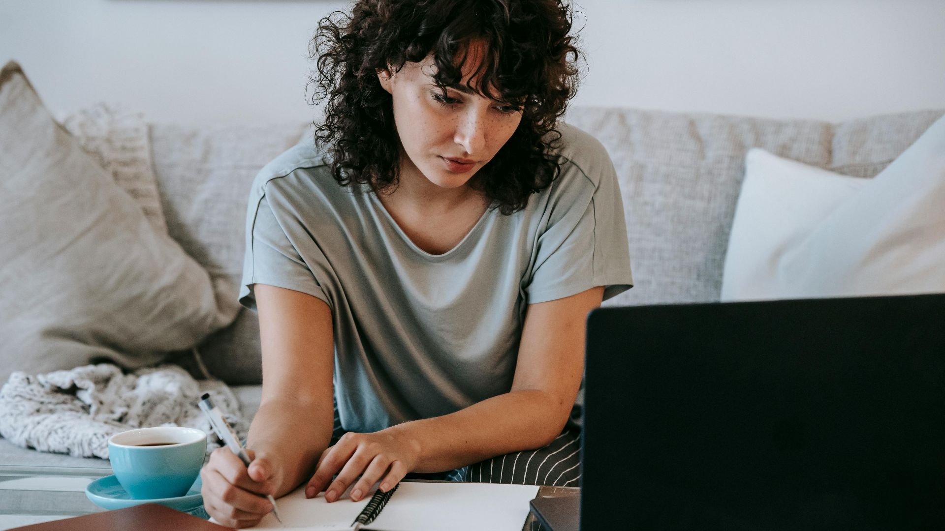 Focused young female student with dark curly hair in casual clothes taking notes in copybook while preparing for exams sitting on couch and using laptop at home
