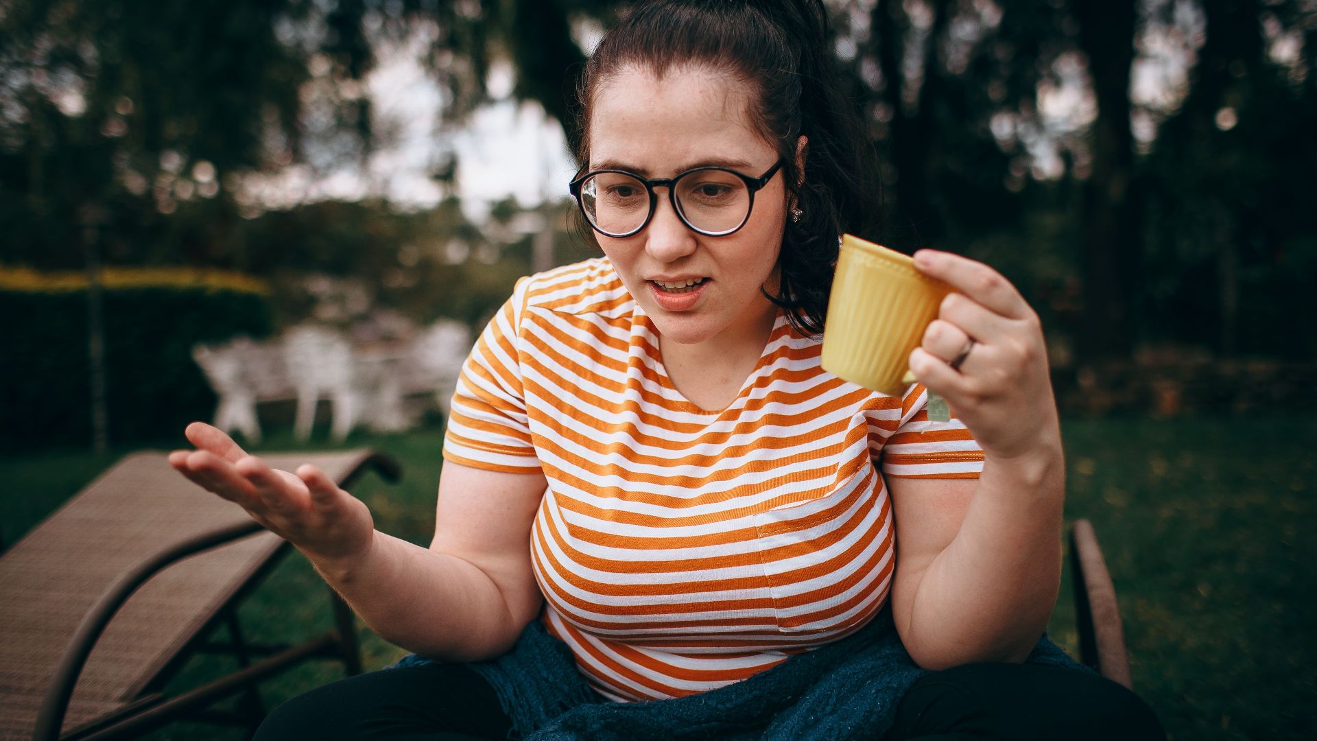 Young woman enjoying a coffee outdoors in a park setting, wearing glasses and a striped shirt.