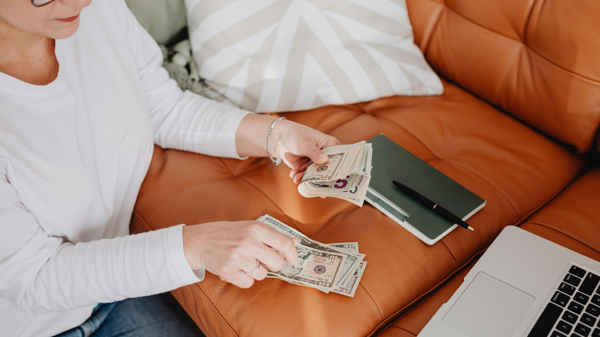 An adult woman sitting on a sofa, counting money with a laptop and notebook nearby, illustrating budget planning.