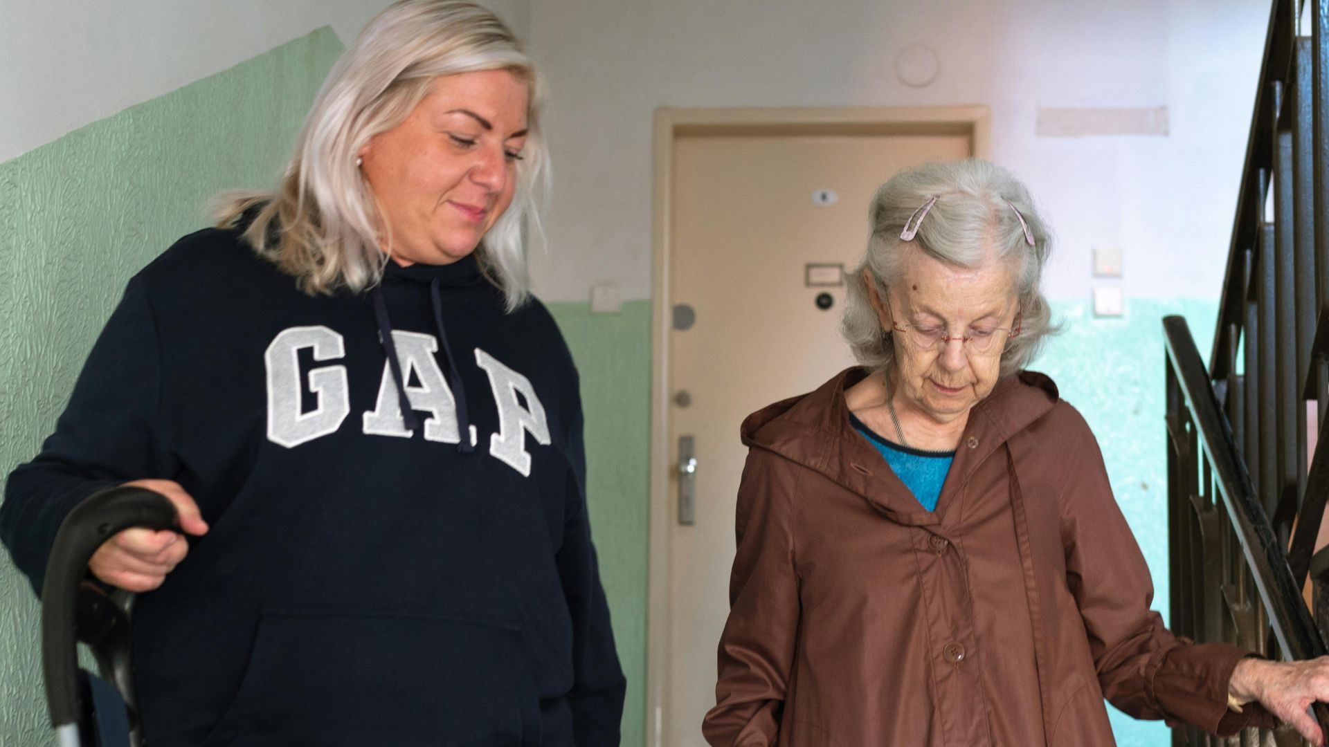 A senior woman assisted by a caregiver walking down indoor stairs in Prague.
