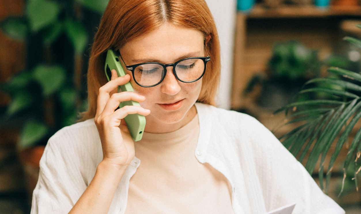 Woman in glasses discussing document on phone surrounded by plants indoors