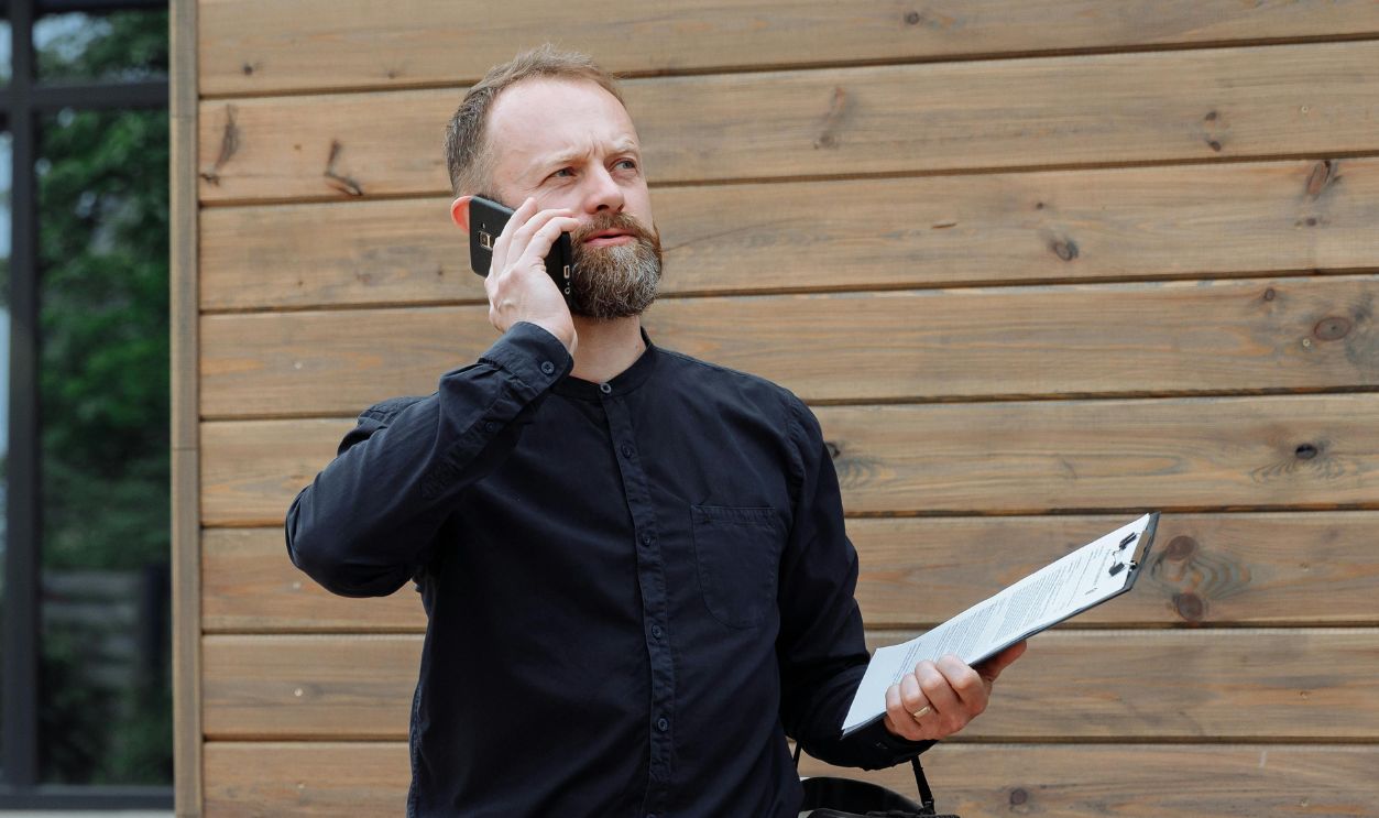 Real estate agent making a phone call while holding documents near home for sale sign