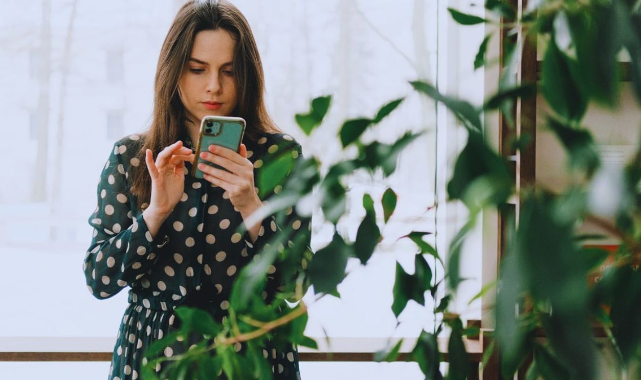 Woman in polka dot dress using smartphone indoors, surrounded by plants