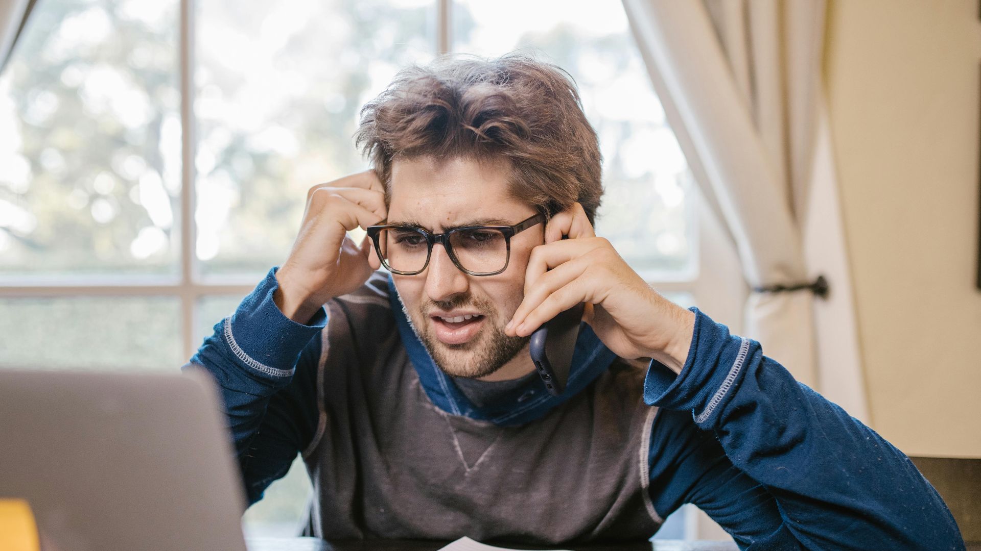 Man in glasses using laptop and smartphone at home, appearing confused.