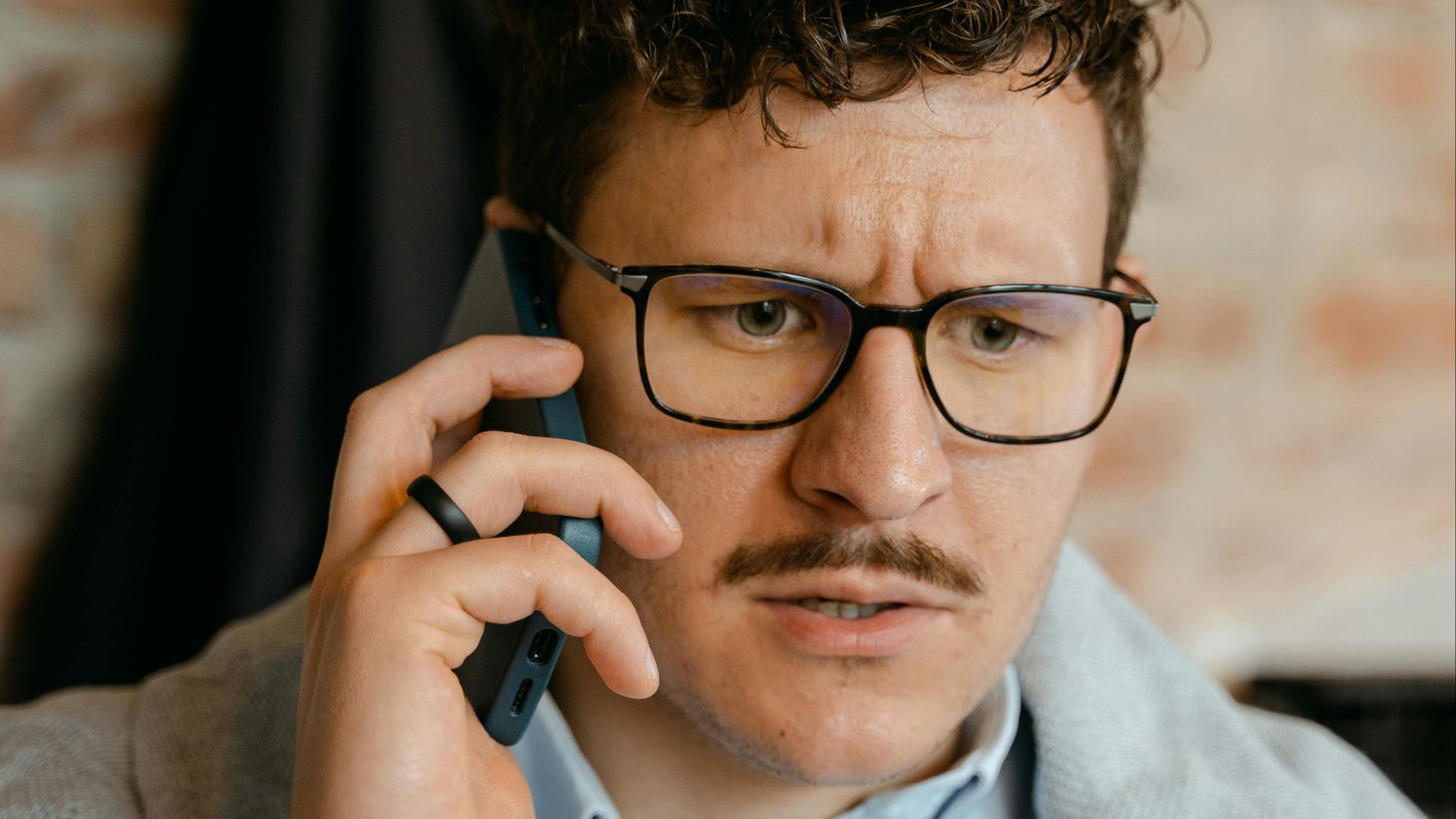 A man with curly hair and glasses engaged in a phone conversation indoors.