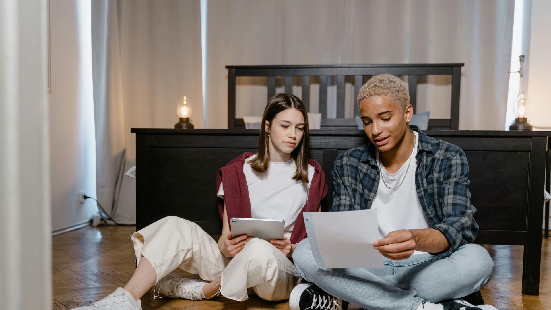 Two young adults studying with a tablet and papers on a bedroom floor.