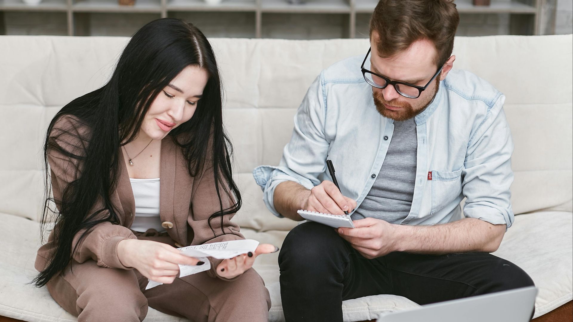 A man and woman sitting indoors, reviewing expenses with notebooks and money on the table.