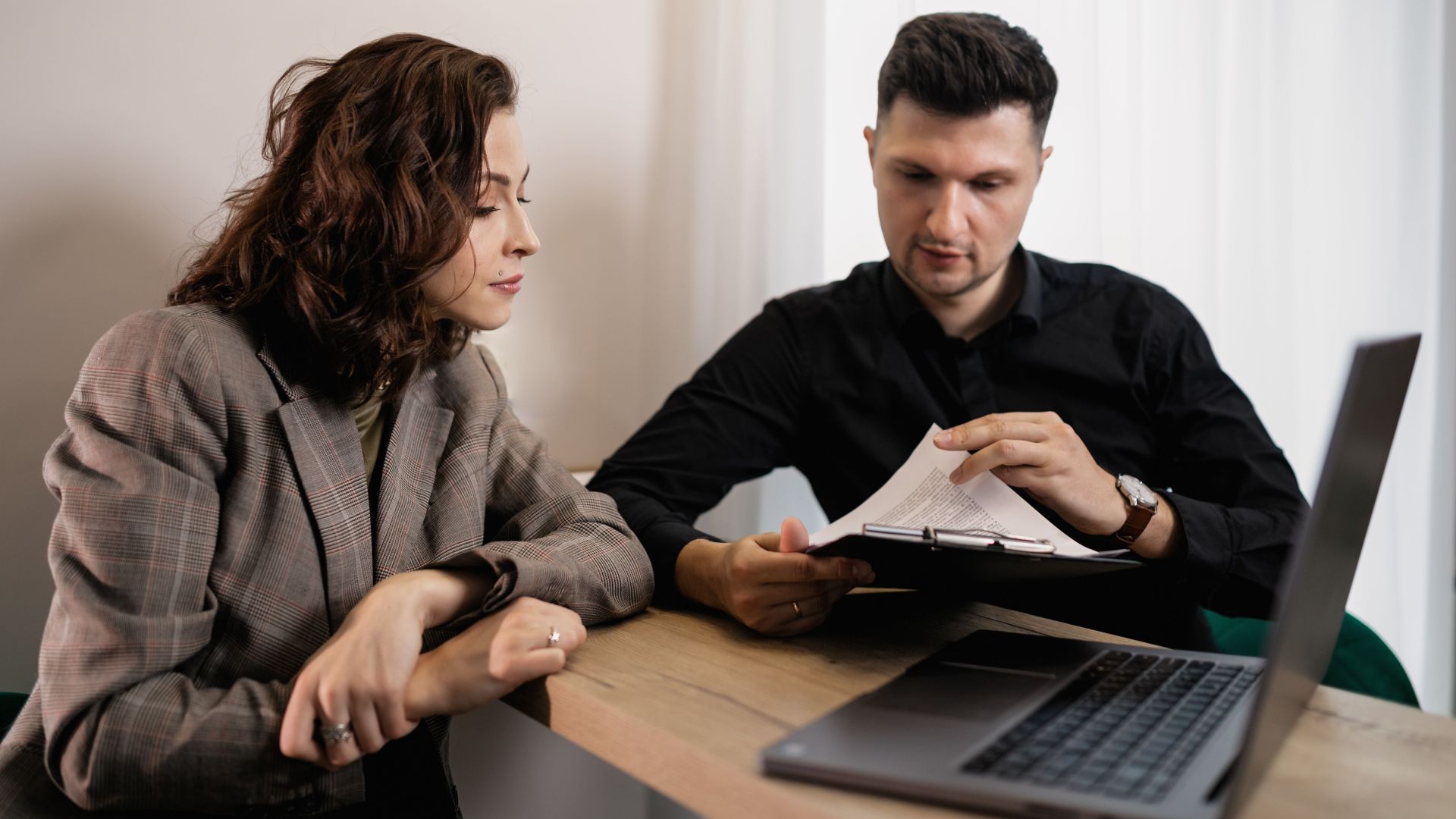 Two professionals discussing documents at a desk with a laptop, focused on collaboration.