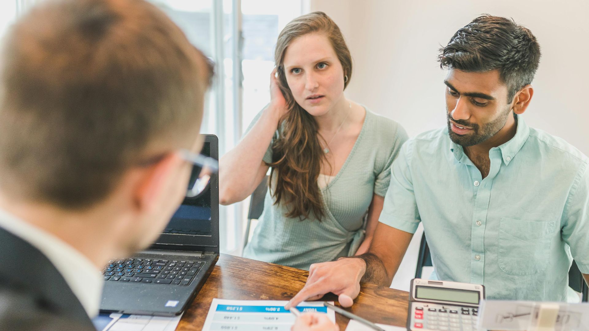 A young couple consults with a realtor about buying a home, indoors in a modern office setting.