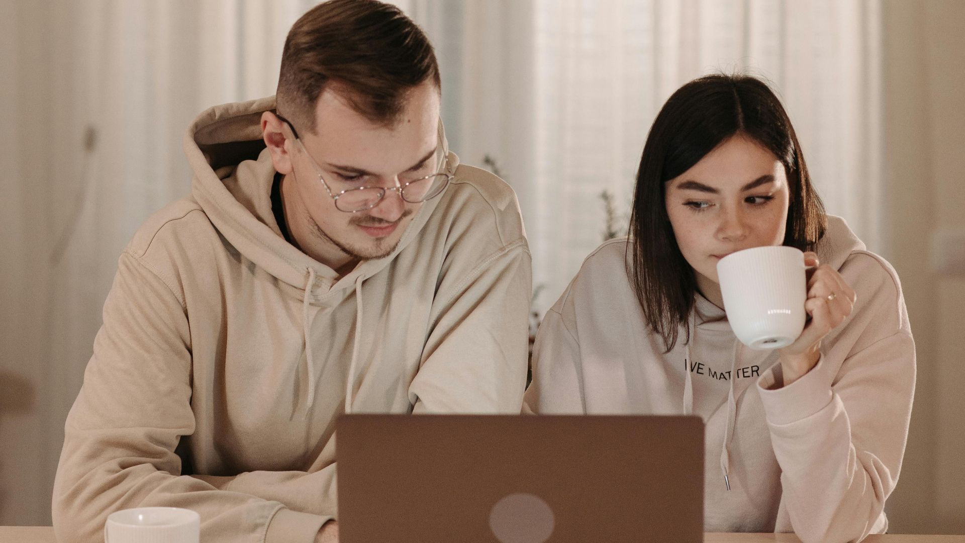 A cozy indoor setting with a couple in hoodies enjoying coffee and working on a laptop.