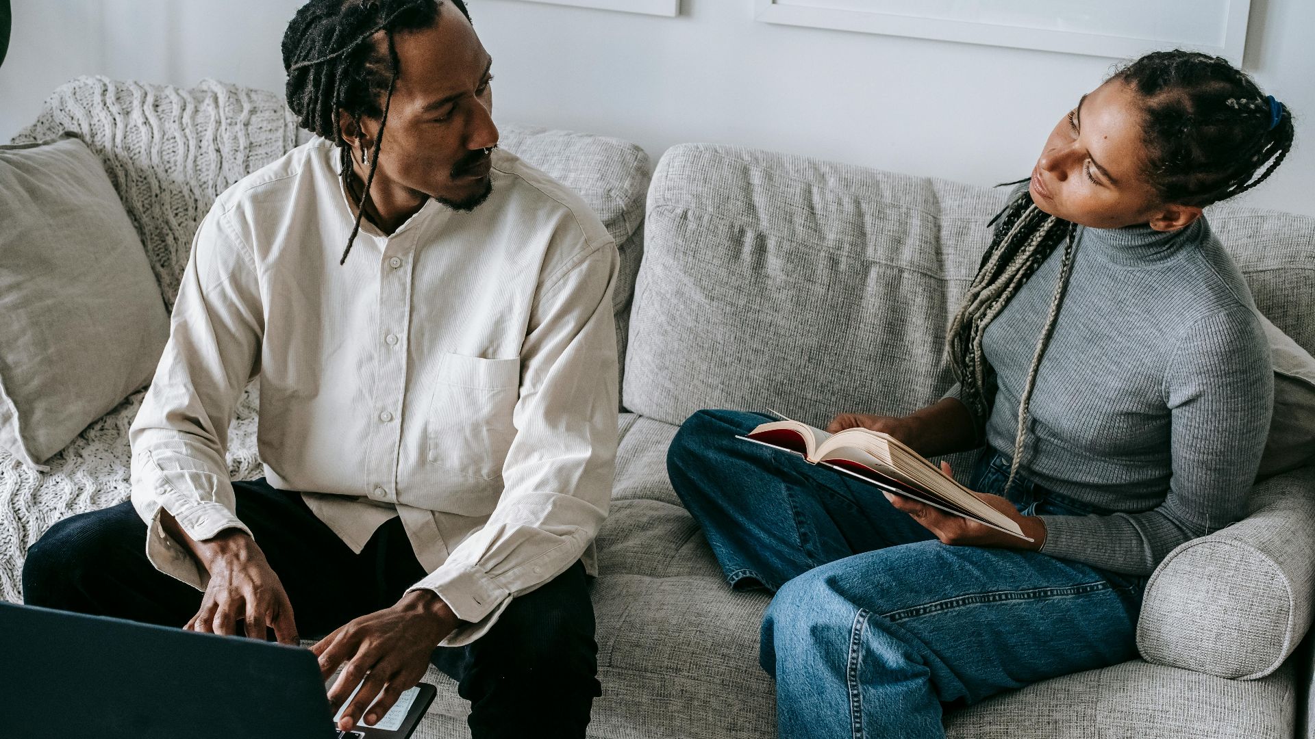 From above of serious young African American husband and wife in casual outfits sitting on sofa with book and laptop and discussing relationships in cozy living room