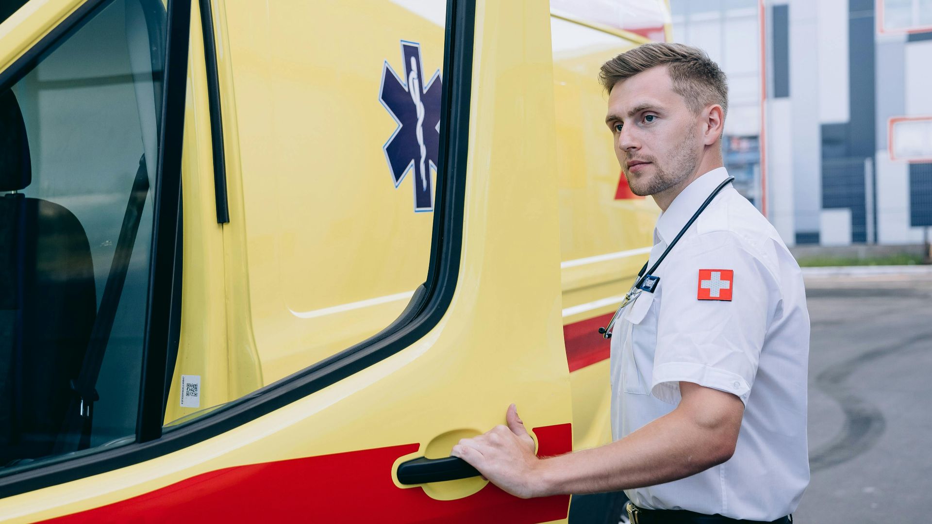 A young male paramedic in uniform opens the door of a yellow ambulance outdoors.