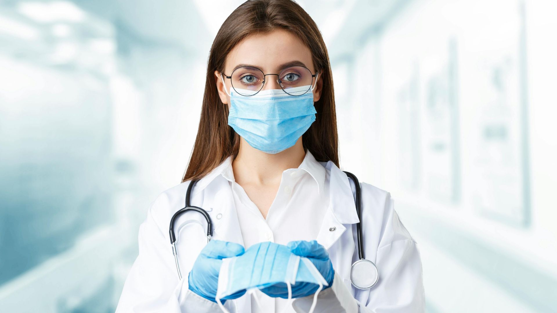 Young female doctor holding a face mask in hospital wearing gloves and stethoscope.
