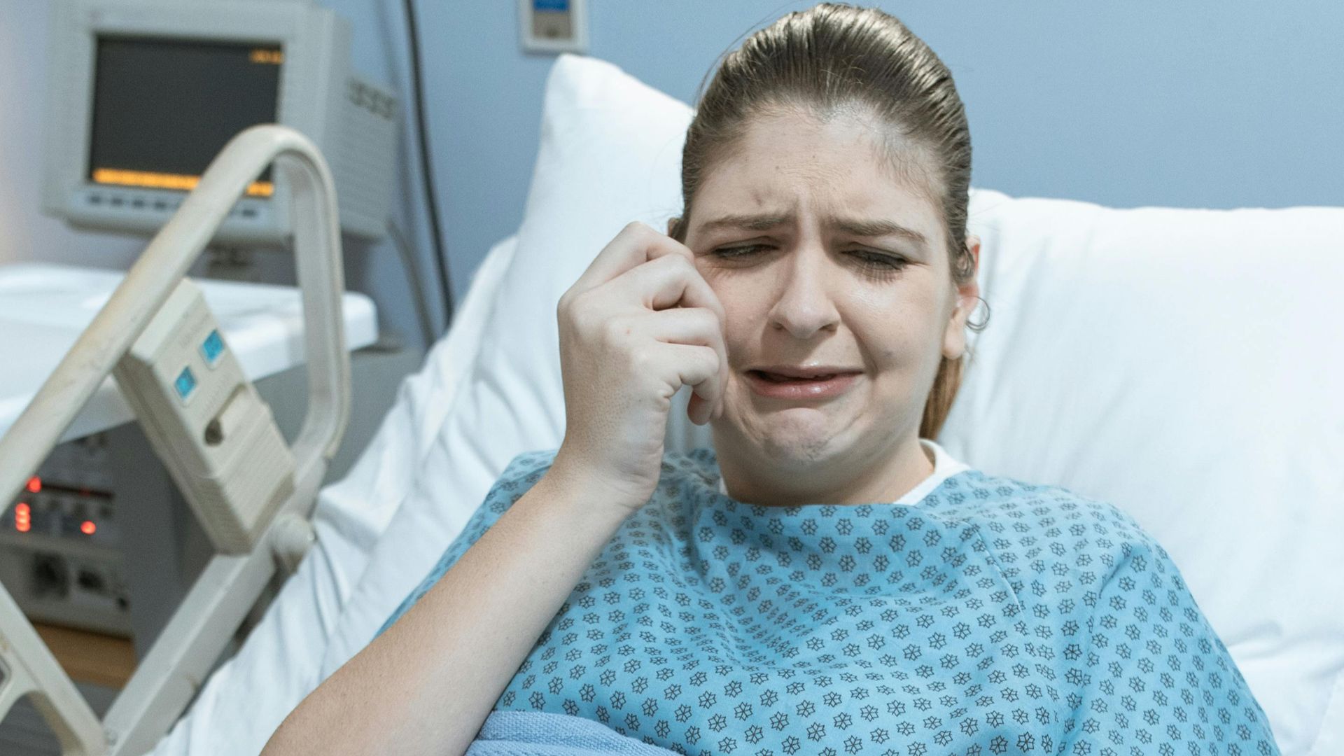 A woman in a hospital bed, comforted by a hand, wearing a gown and blanket.