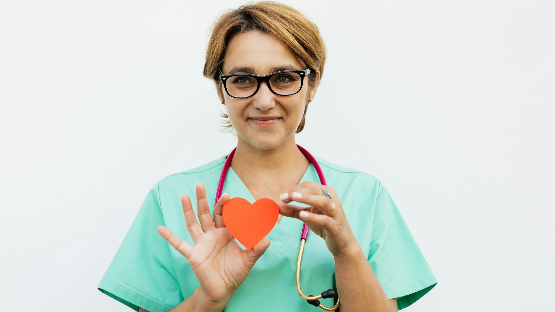 Female doctor in scrubs holding a paper heart symbol, smiling with a stethoscope around her neck.