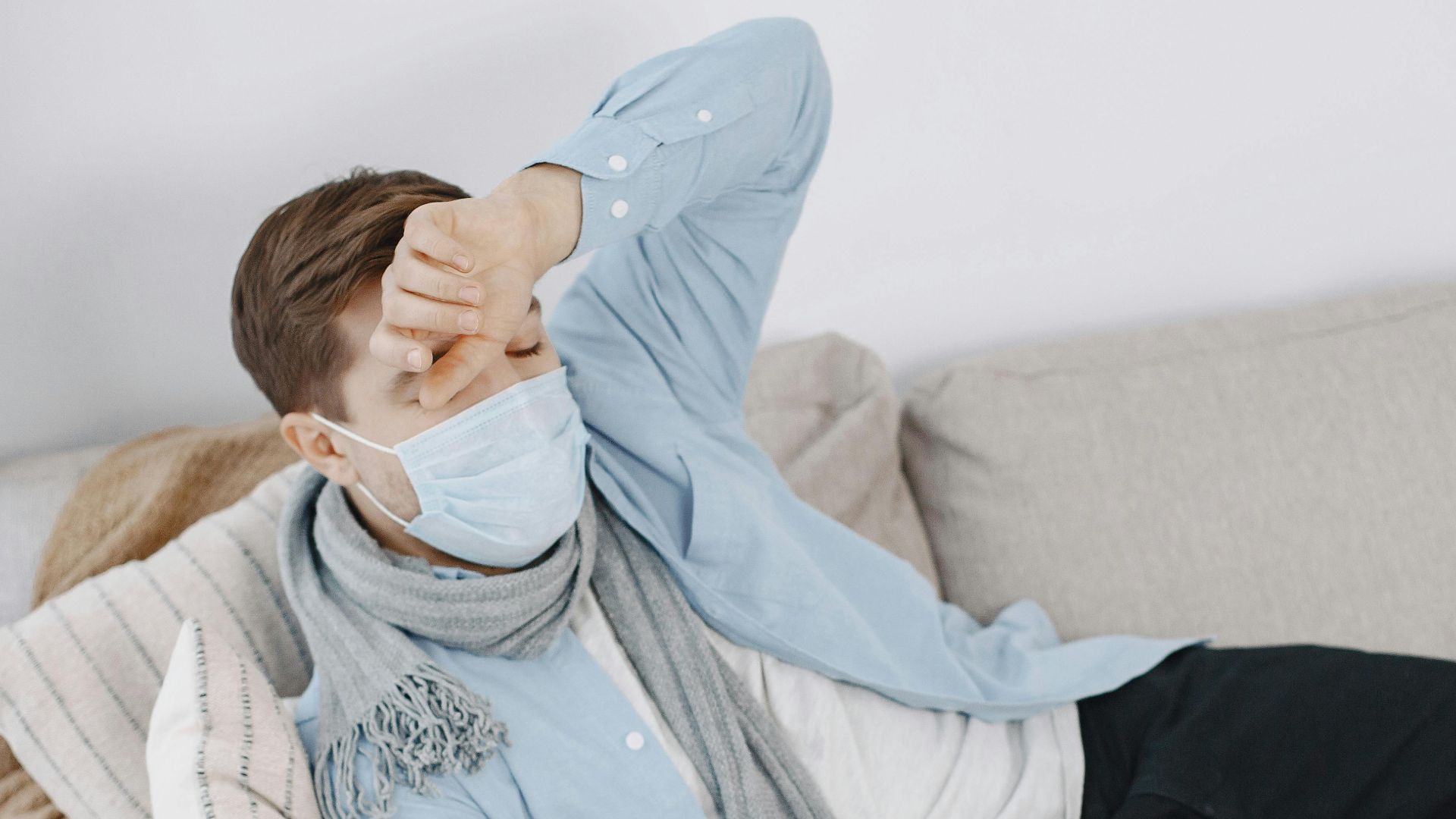 Man in mask resting on sofa with pulse oximeter and tablet, conveying illness at home.