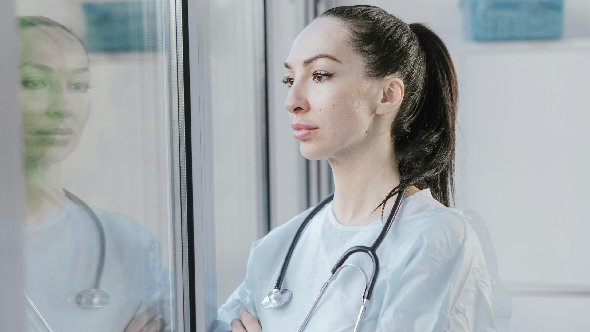 Female doctor in a lab coat with stethoscope, pondering while looking out a window.