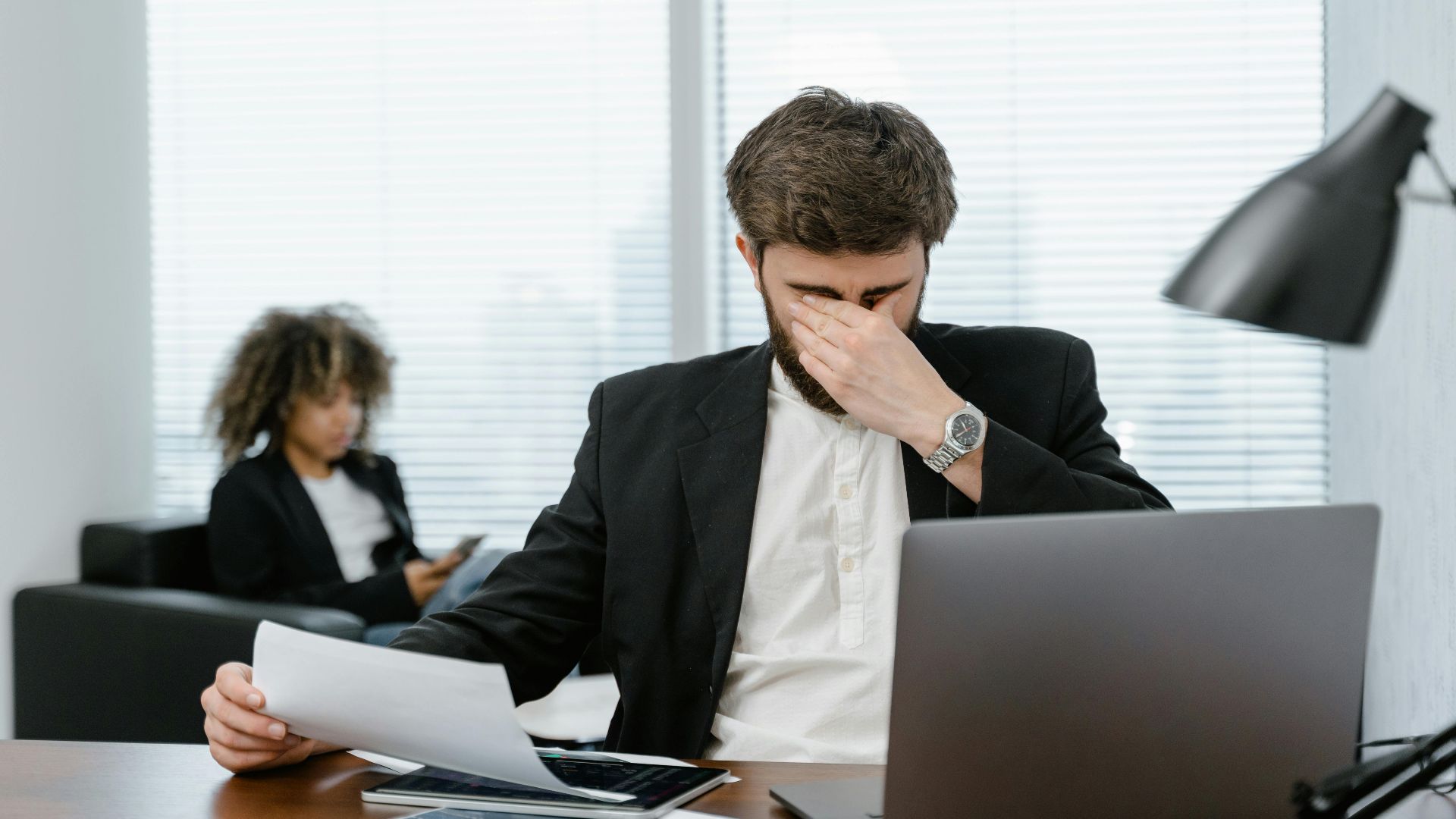 Businessman in an office covering his eyes while working on a laptop, feeling stressed.