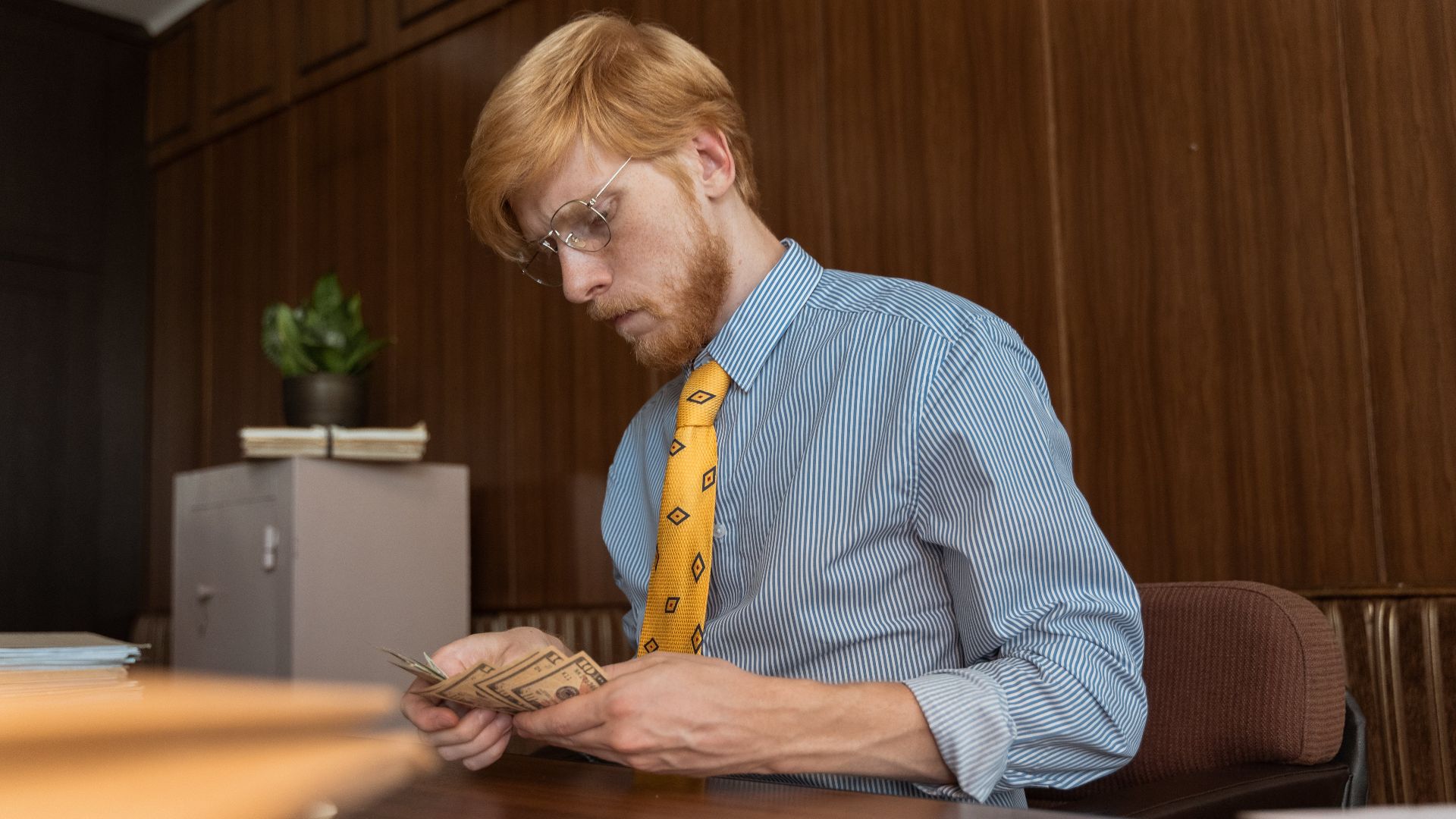 Serious businessman counting cash at his desk in an office environment with wood paneling.