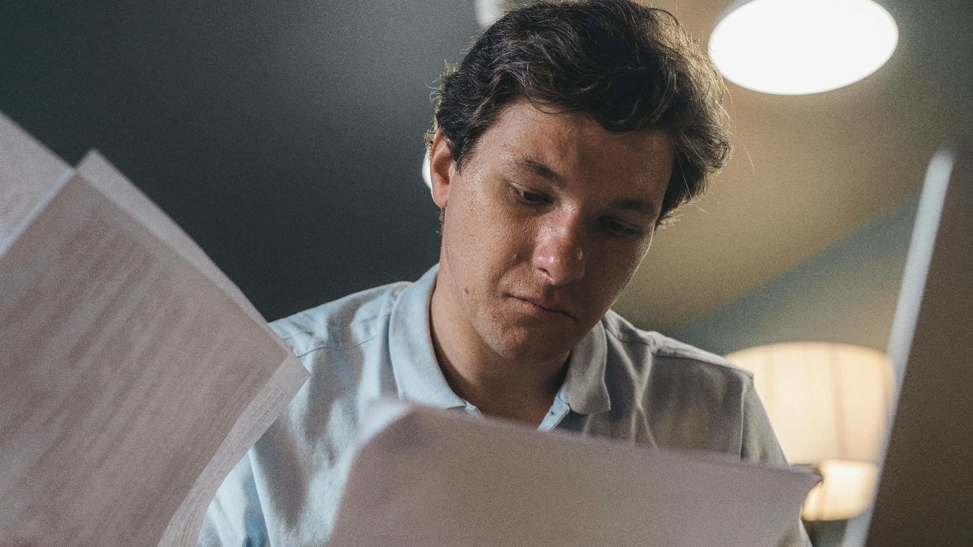 Serious man wearing a light blue shirt intently reading printed papers indoors under warm lighting.
