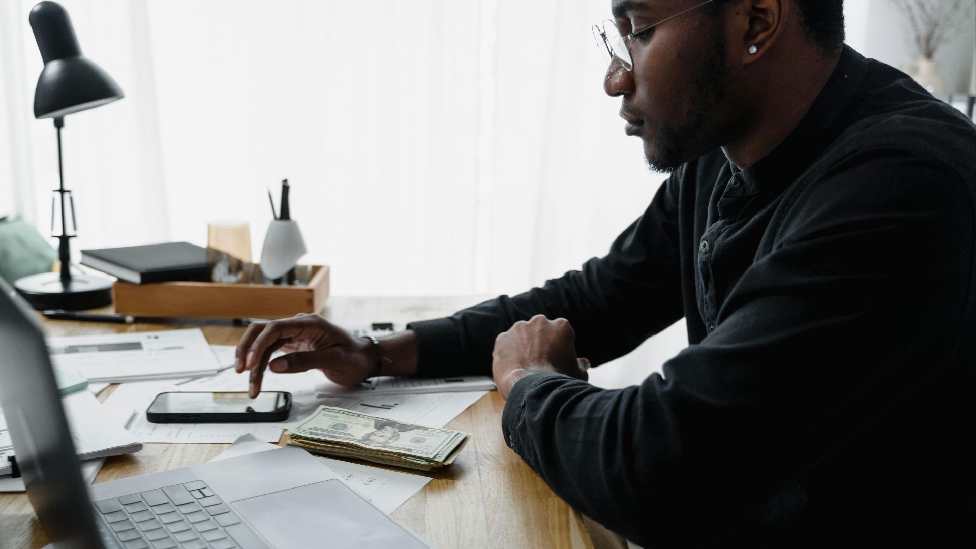 Businessman in office using smartphone for budgeting with cash and laptop on desk.