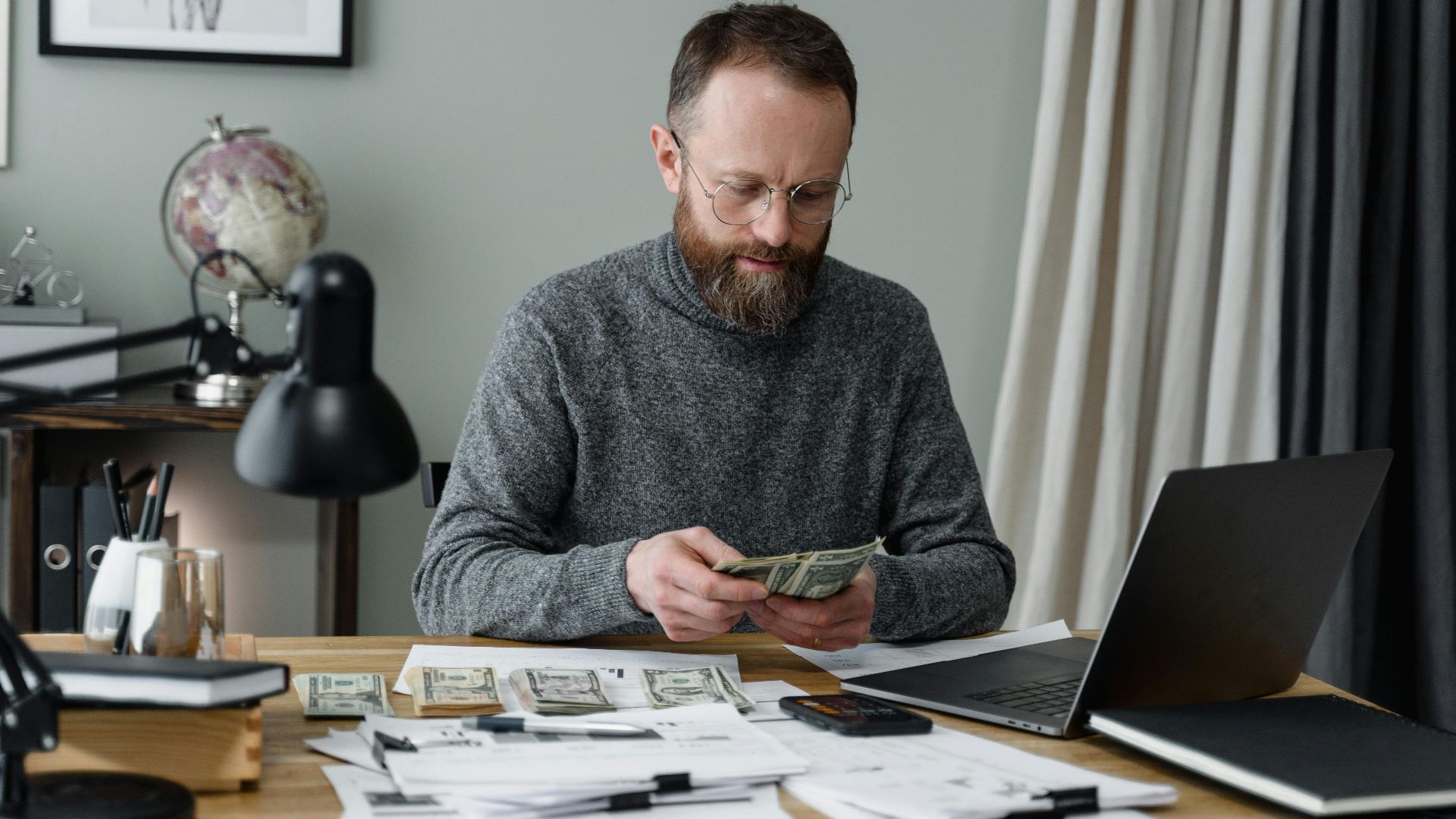 Businessman counting cash at a desk with documents and a laptop in an office.