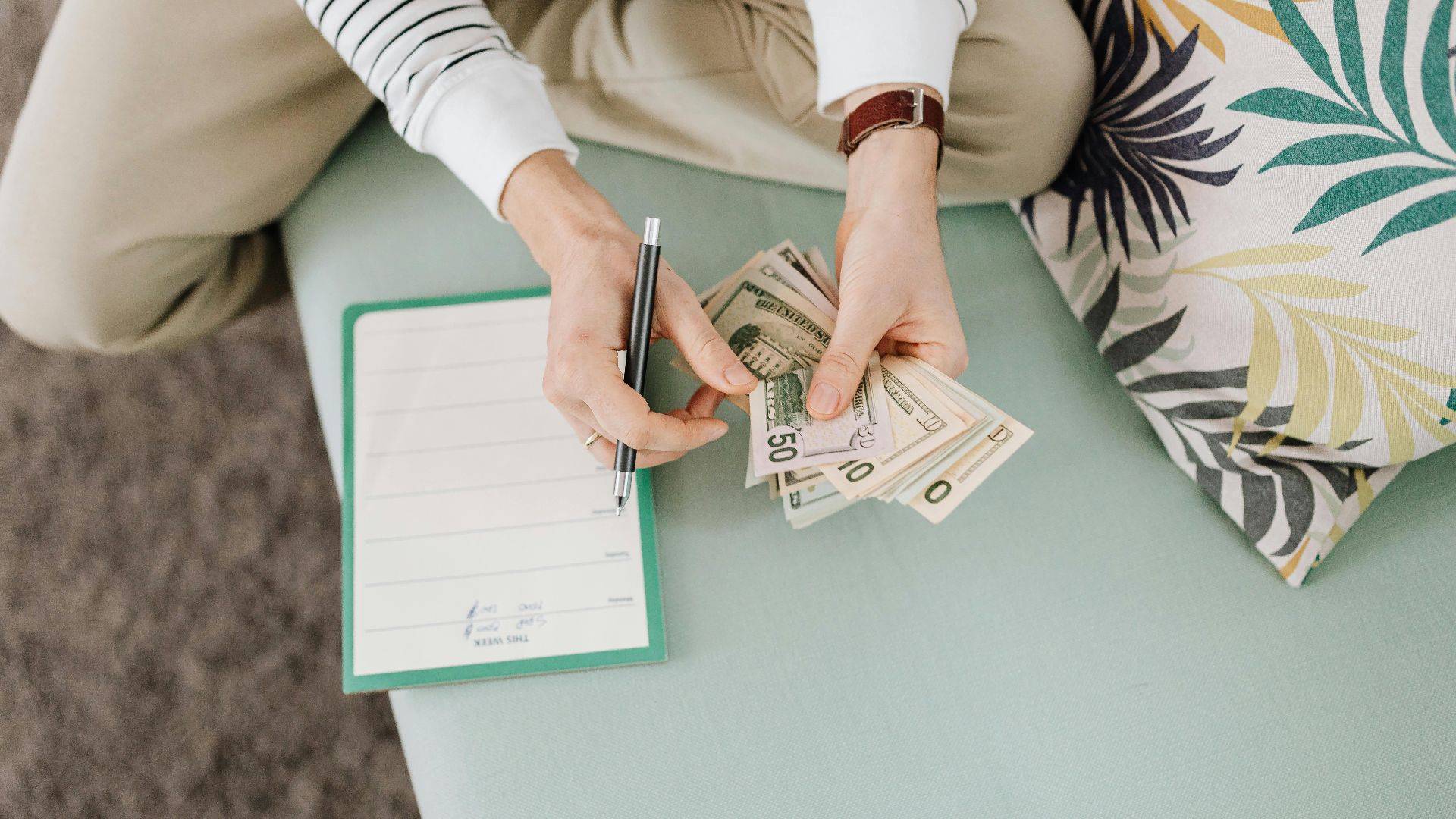 Person holding US dollar bills with a notepad and pen on a soft surface indoors.