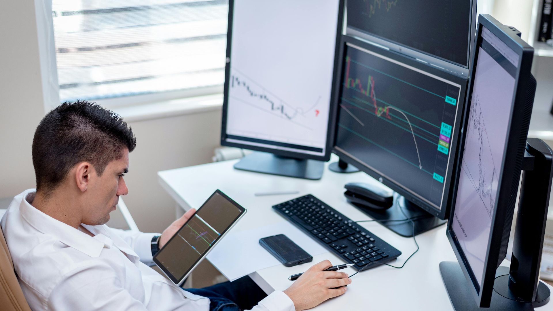 Business professional analyzing financial charts on monitors and tablet in modern office workspace.