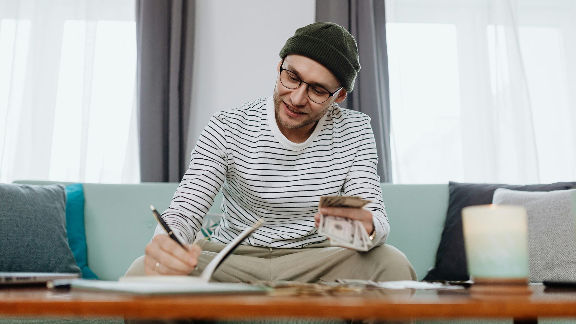 Smiling man in beanie counts cash while writing at home.