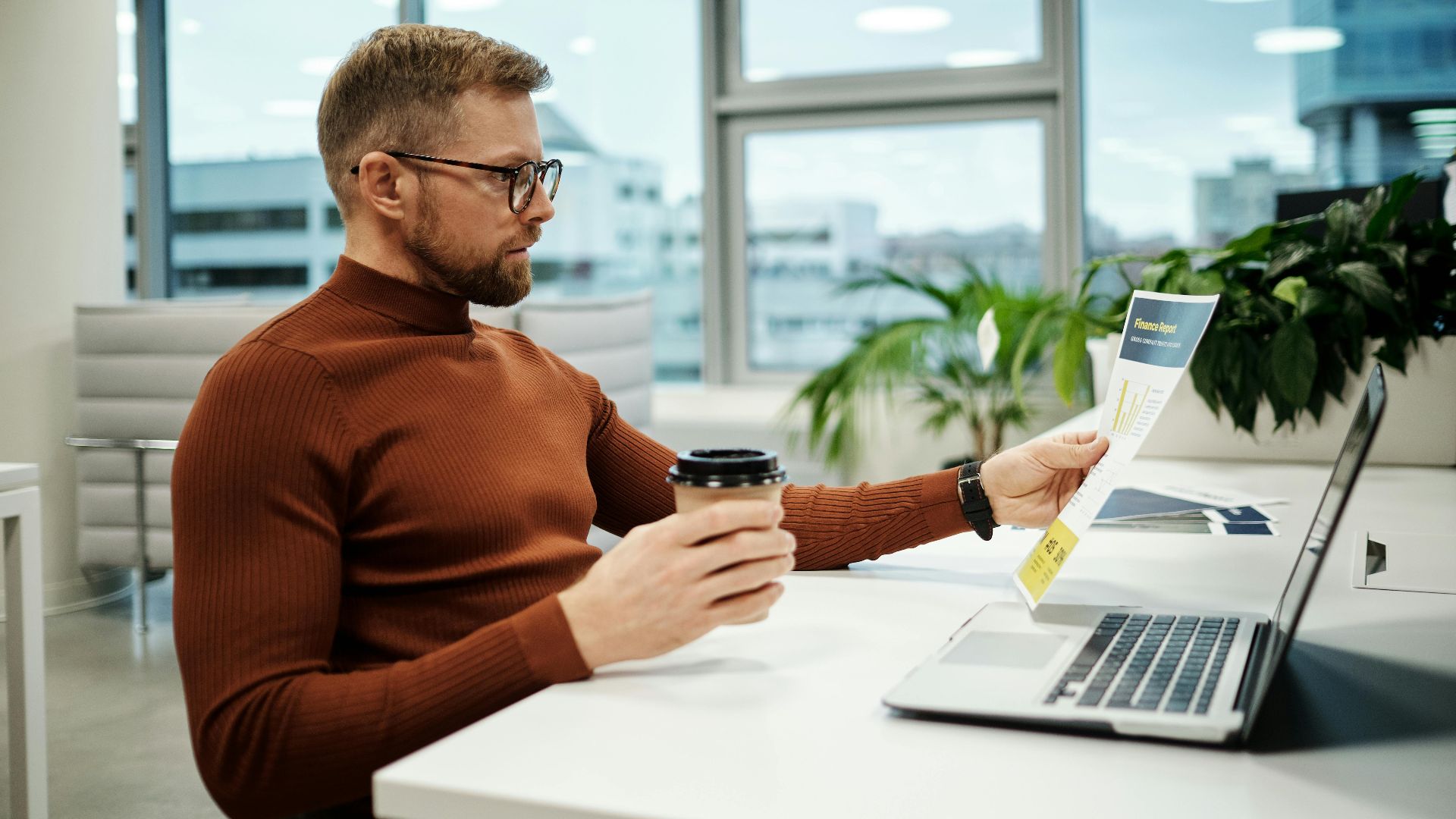 Professional businessman in brown turtleneck analyzing financial report while holding a takeaway coffee in modern office.
