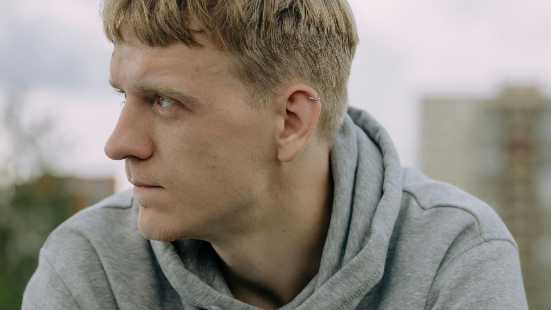 A young man in a gray hoodie looking thoughtfully into the distance outdoors.