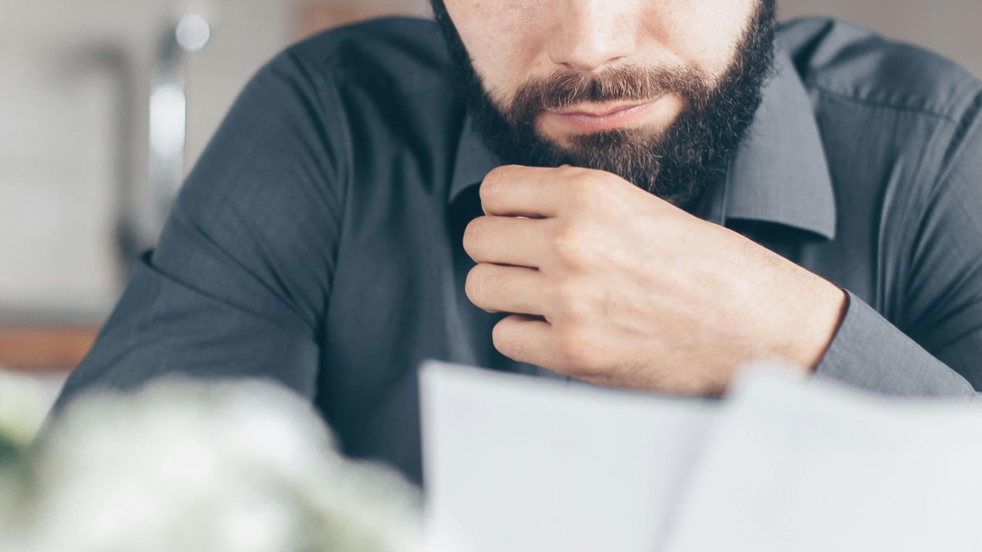A bearded man in a gray shirt intently reads documents while seated indoors, appearing concerned.
