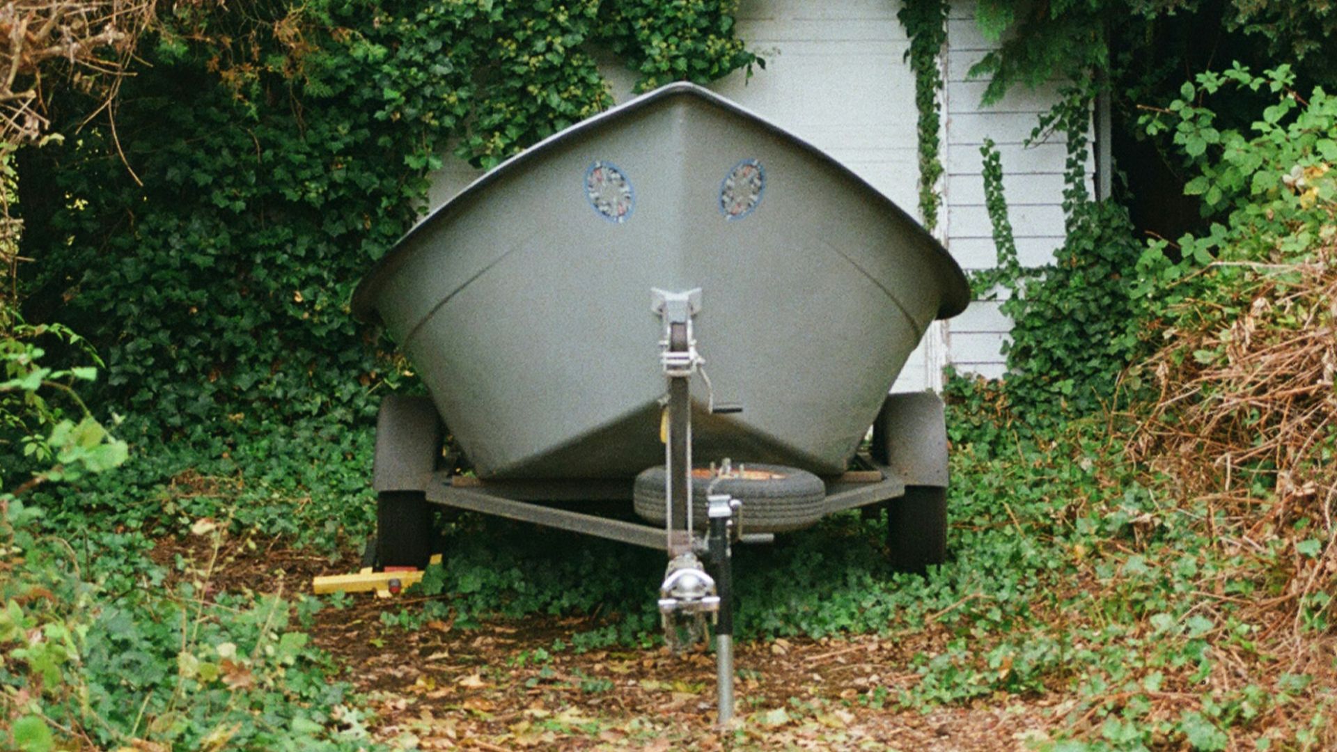 A gray boat sits on a trailer by a shed.