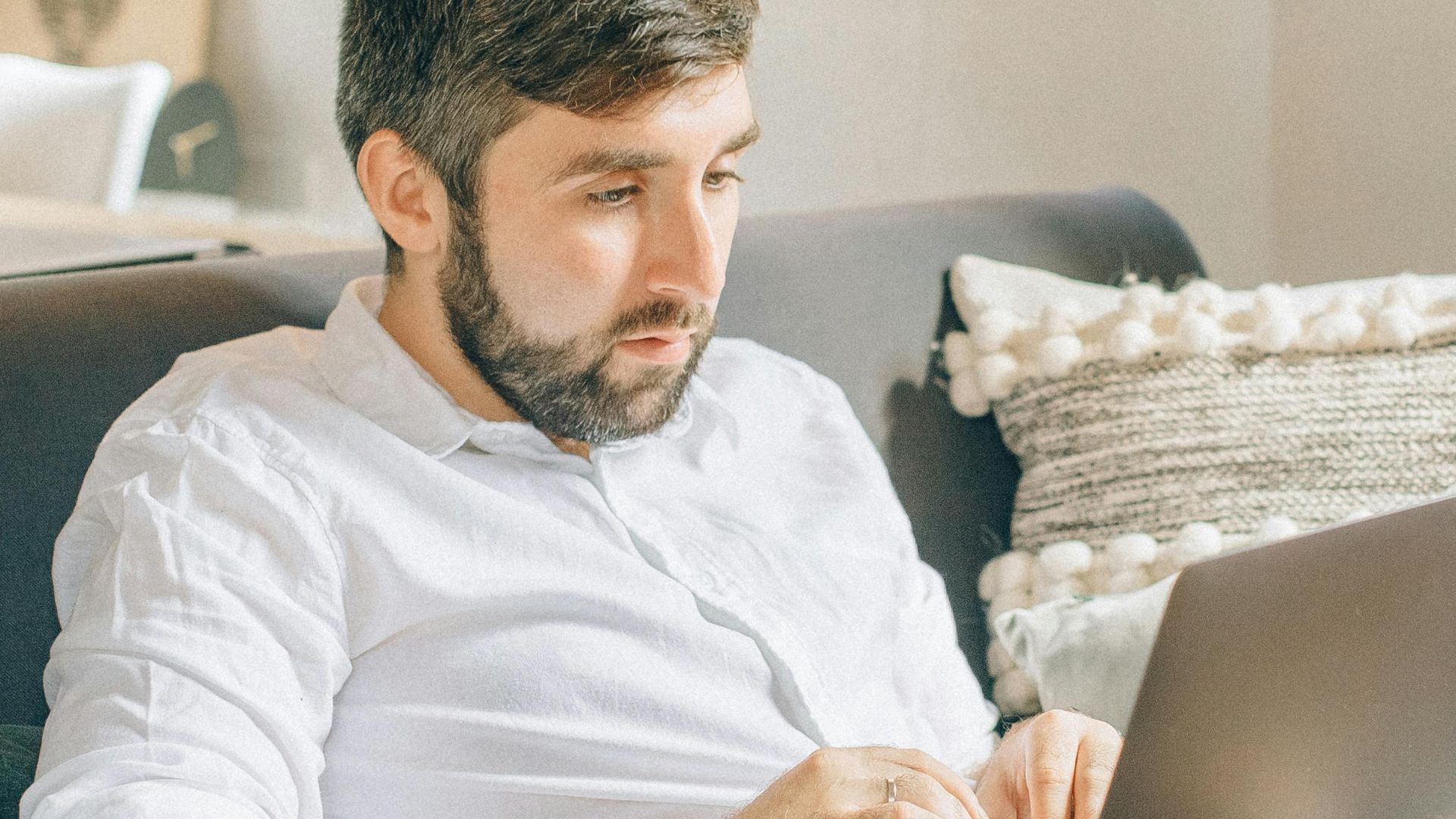 Adult man sitting on couch using laptop, working remotely from cozy home setting.