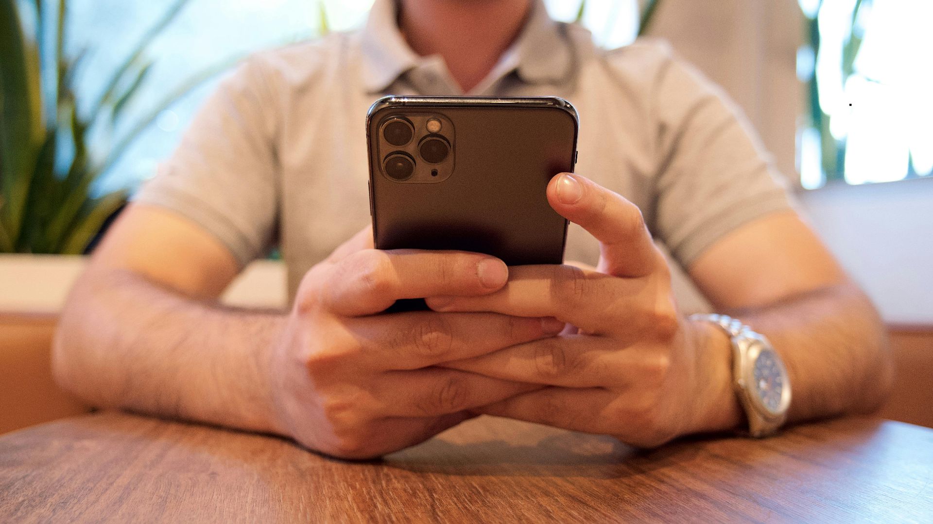Close-up of a man holding a smartphone at a cafe table. Casual setting with plants in the background.