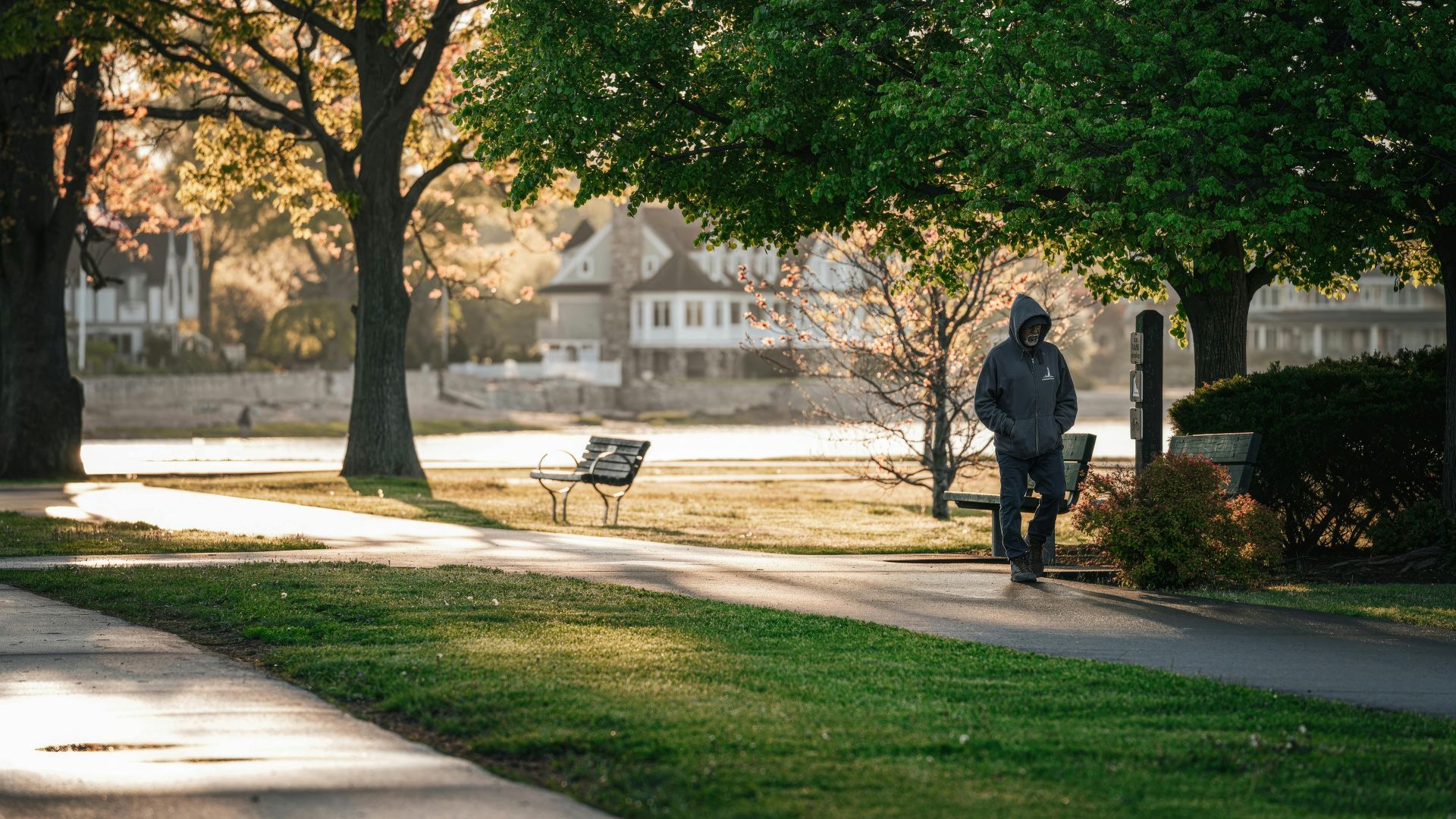 A lone man walks through a lush, green park during daytime, surrounded by trees and sunlight.