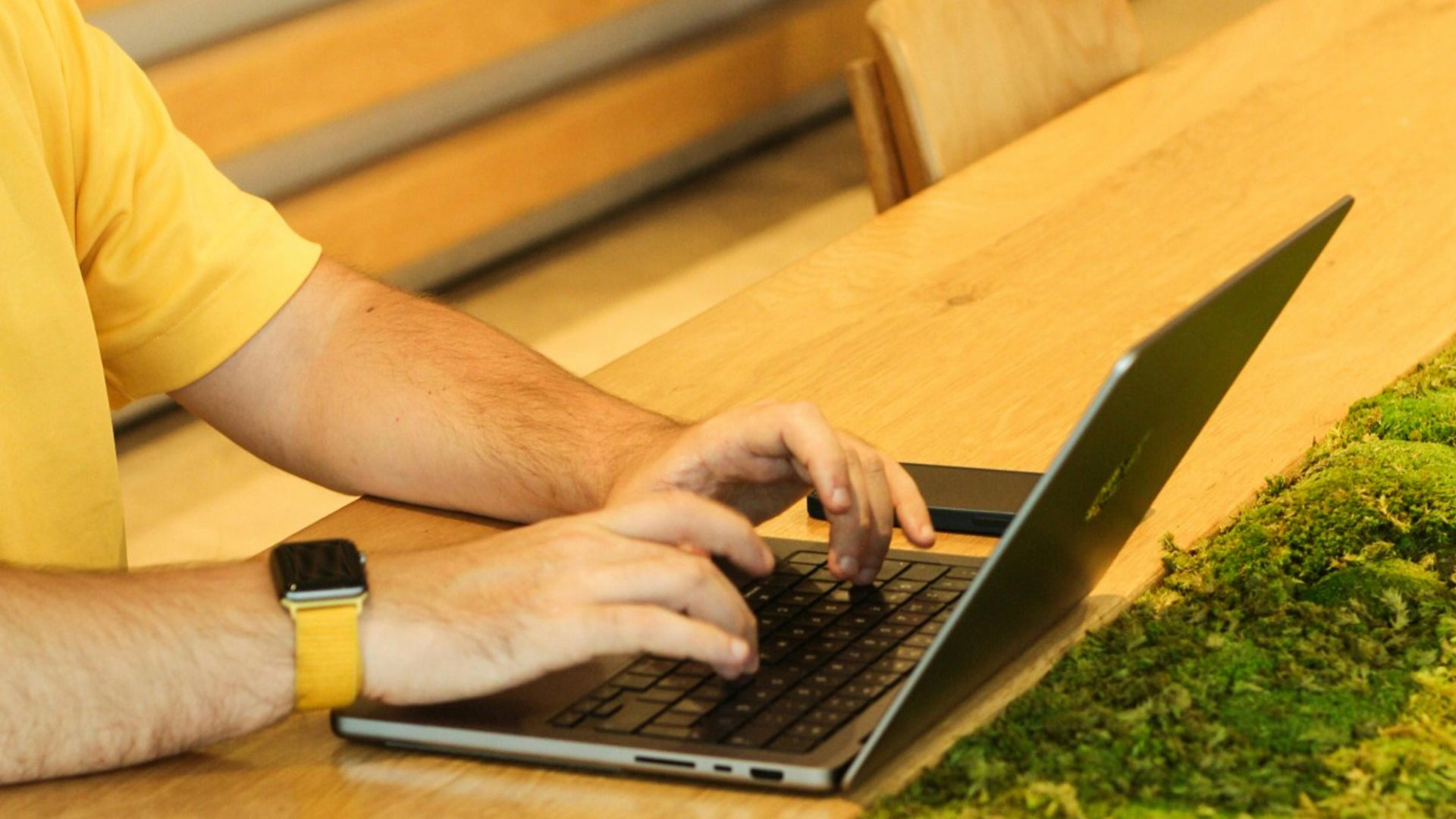 A person using a laptop at a wooden table with moss in a modern cafeteria setting.