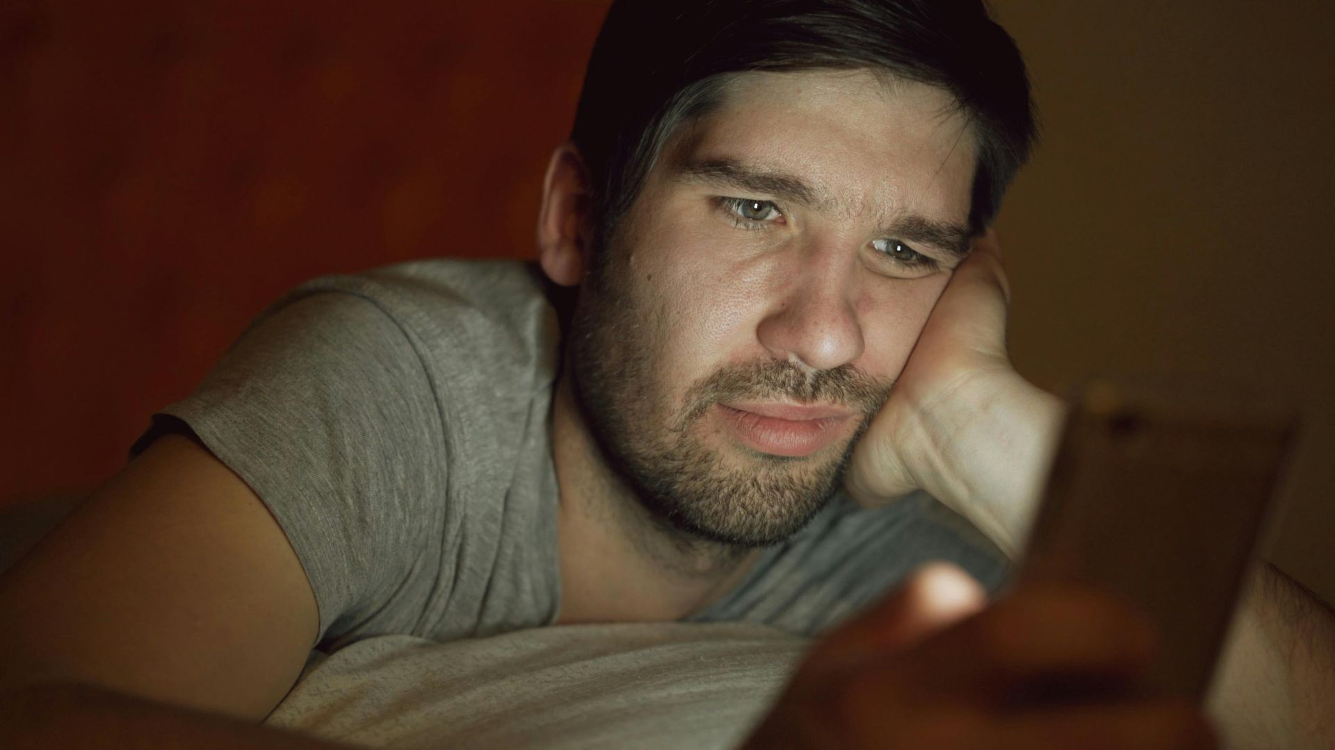 An adult man in a dim room looking at a smartphone with a thoughtful expression.