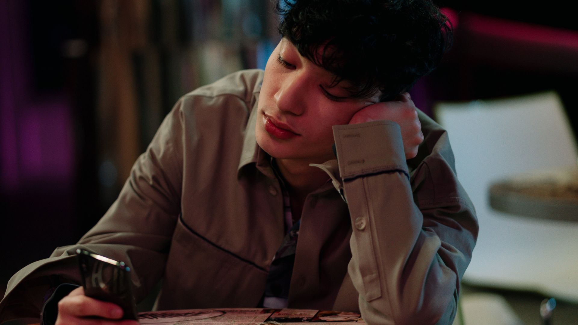 A young man sits indoors looking at his smartphone, appearing thoughtful and relaxed.