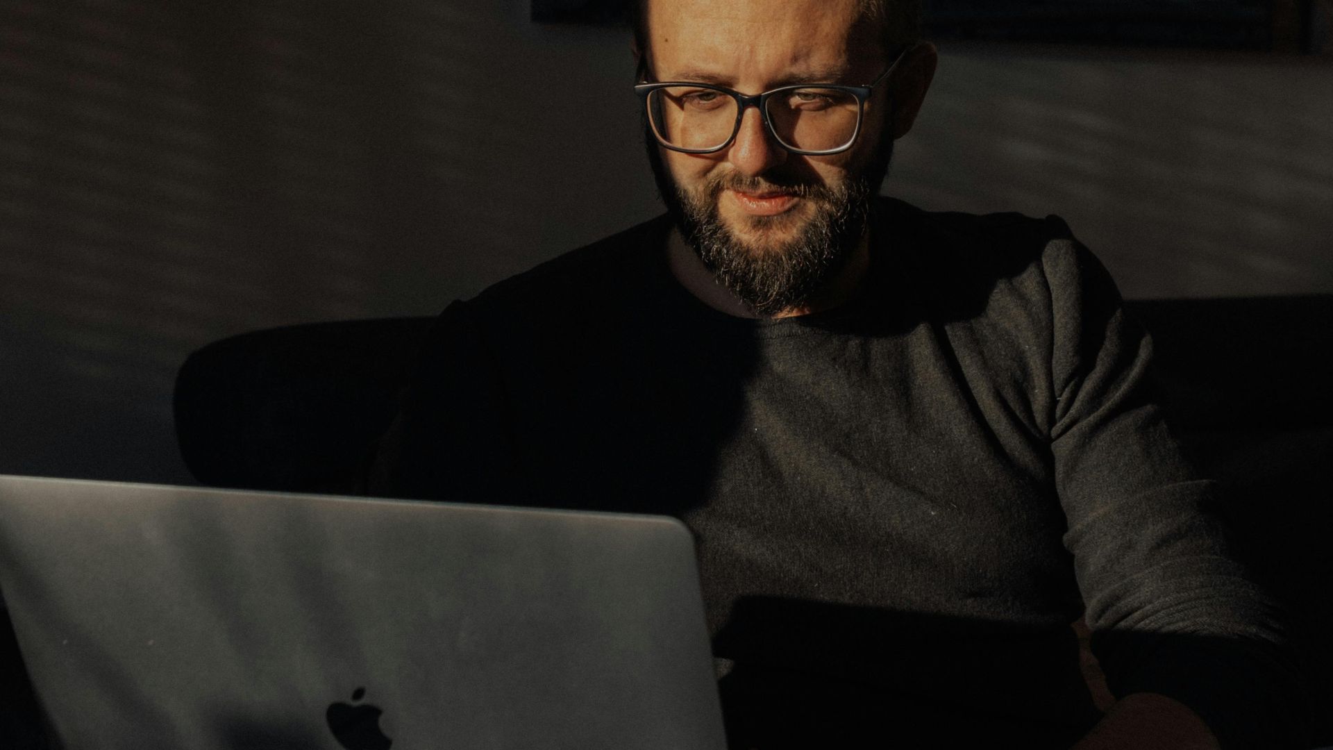 A man with eyeglasses works on a laptop in a dimly lit room, emphasizing productivity and focus.