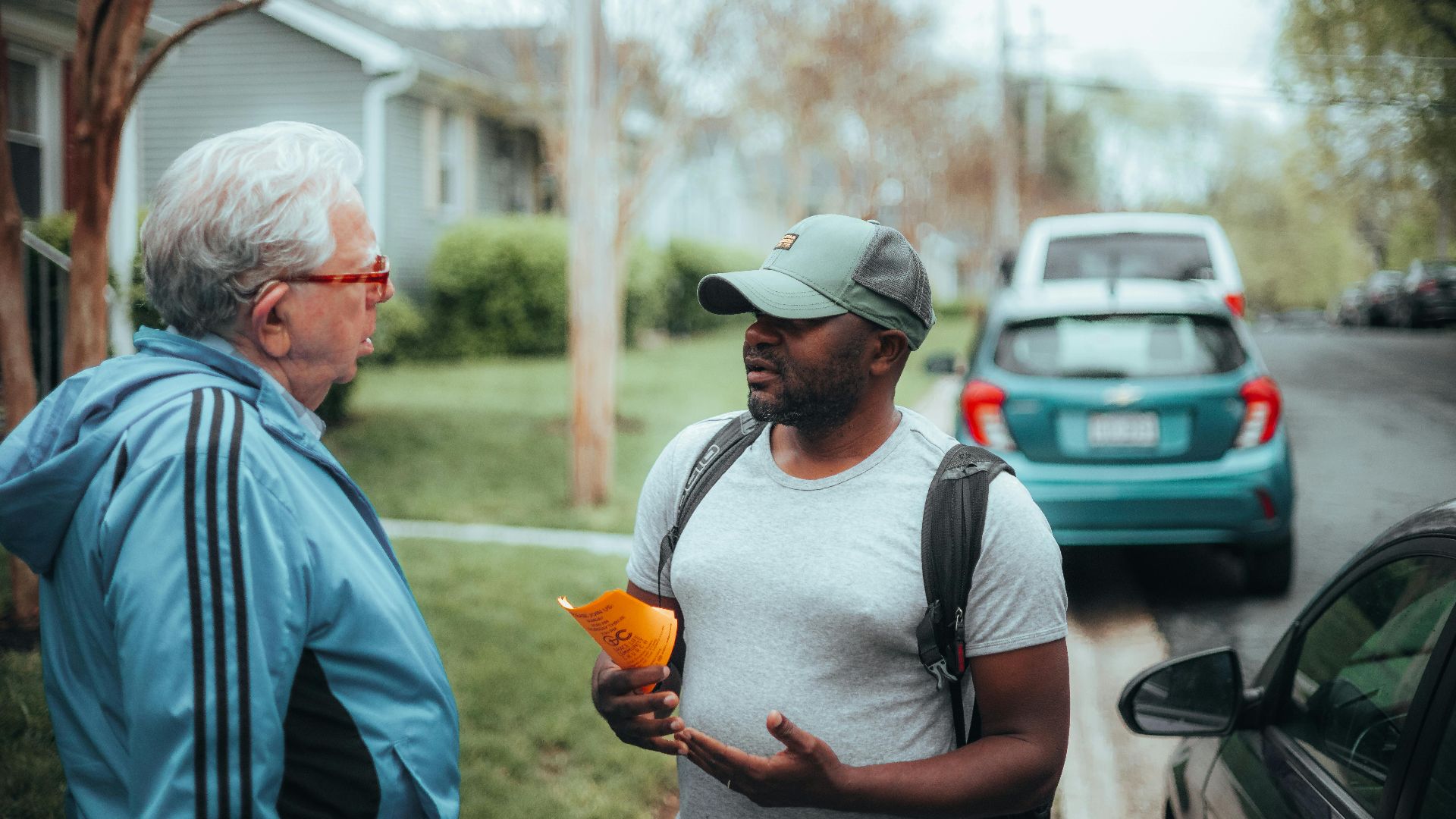 Two men engaged in conversation on a suburban sidewalk with parked cars.