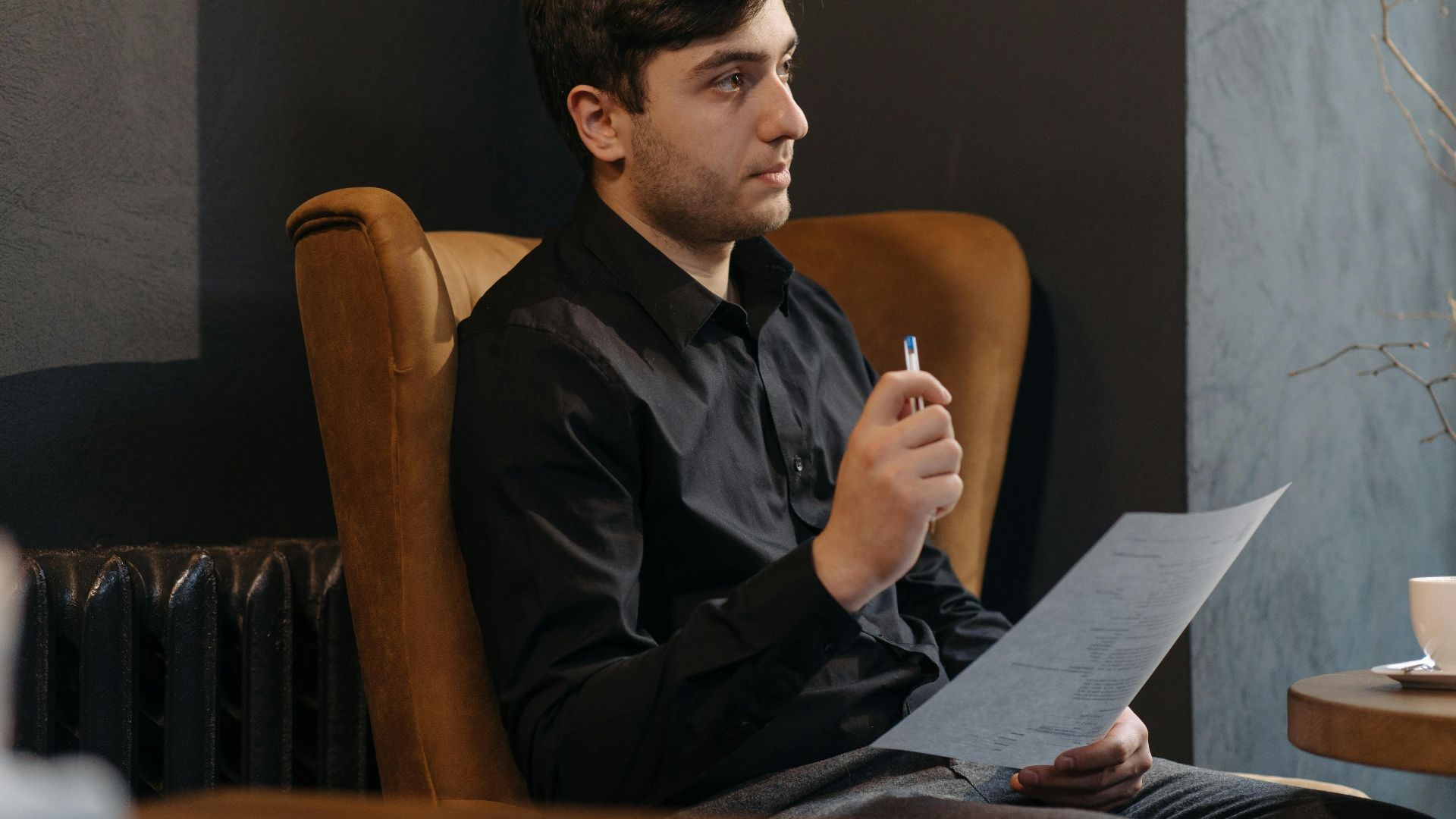Young man in a black shirt sitting thoughtfully with a document and pen indoors.