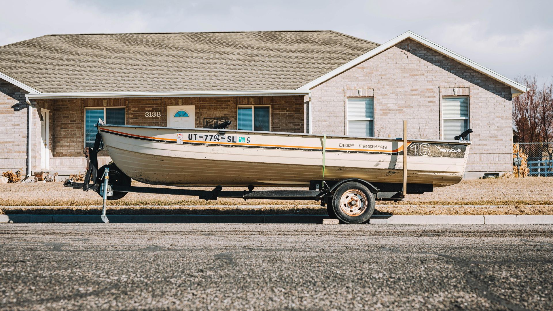 A fishing boat on a trailer parked in front of a suburban house on a clear day.