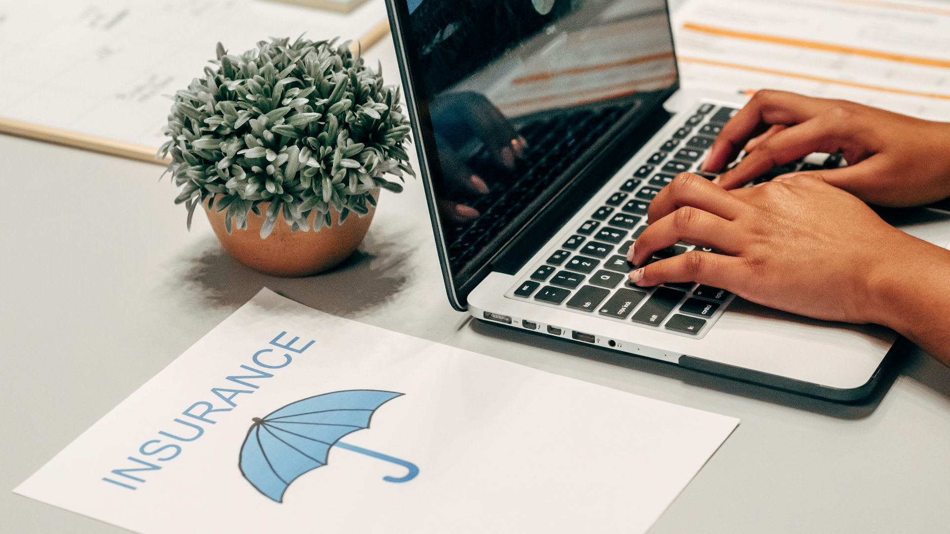 Hands typing on a laptop at a desk with an insurance paper and plant, suggesting a work environment.