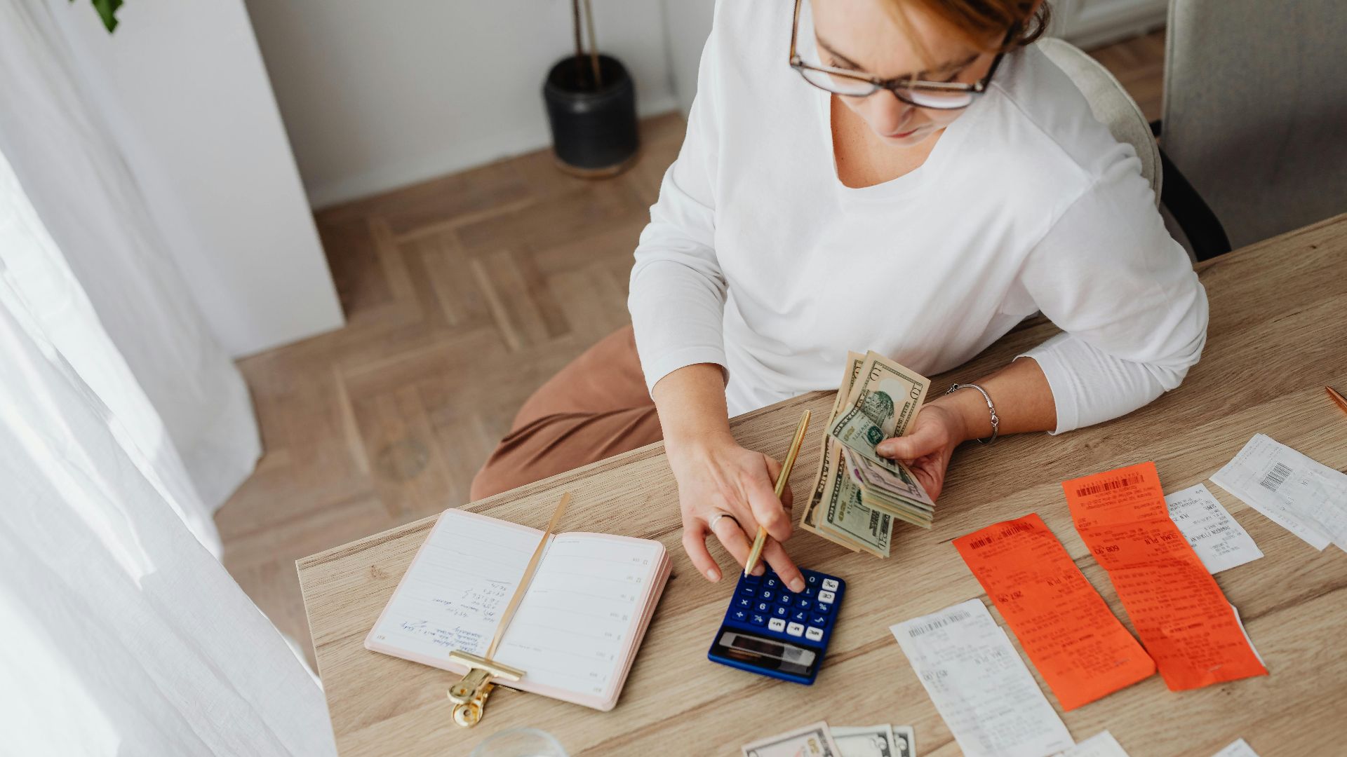 Top view of a woman managing finances with a calculator, cash, and bills in a home setting.
