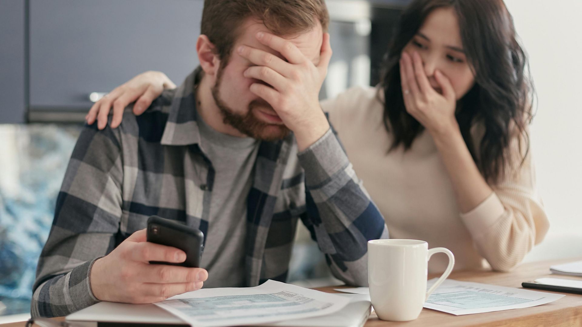 A couple looking stressed and holding a cellphone while reviewing financial documents indoors. Hands on faces in concern.