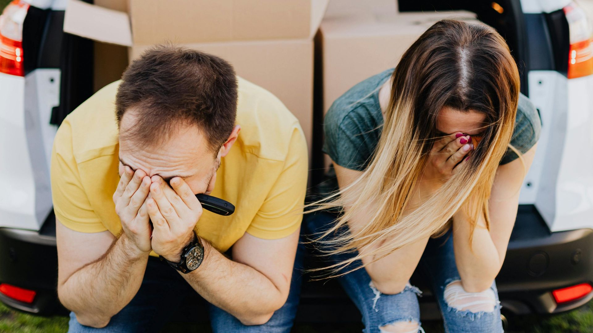 Faceless struggling dissatisfied couple in casual wear sitting on car trunk filled with carton boxed and covering faces with hands after long tiring relocation day