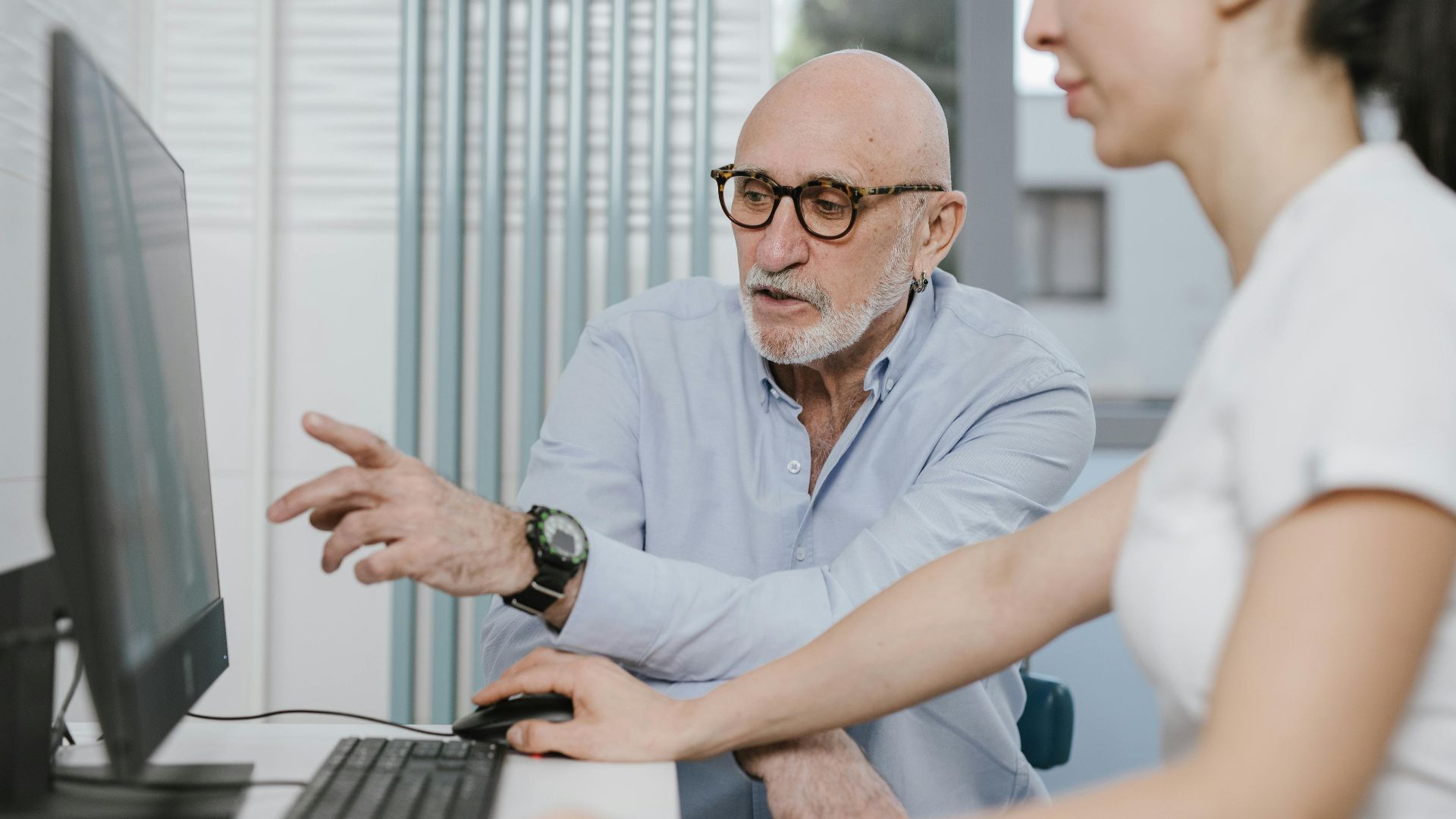 An elderly man pointing at a computer screen while assisting a woman in an office setting.