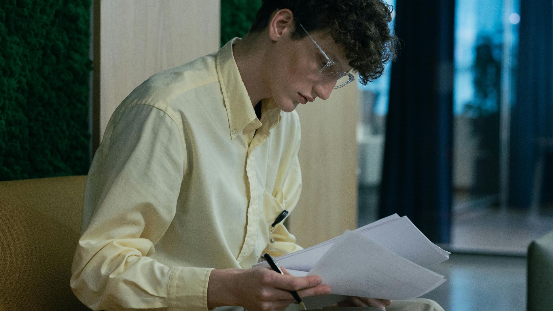 A focused young professional reading documents on a couch in a modern office environment.